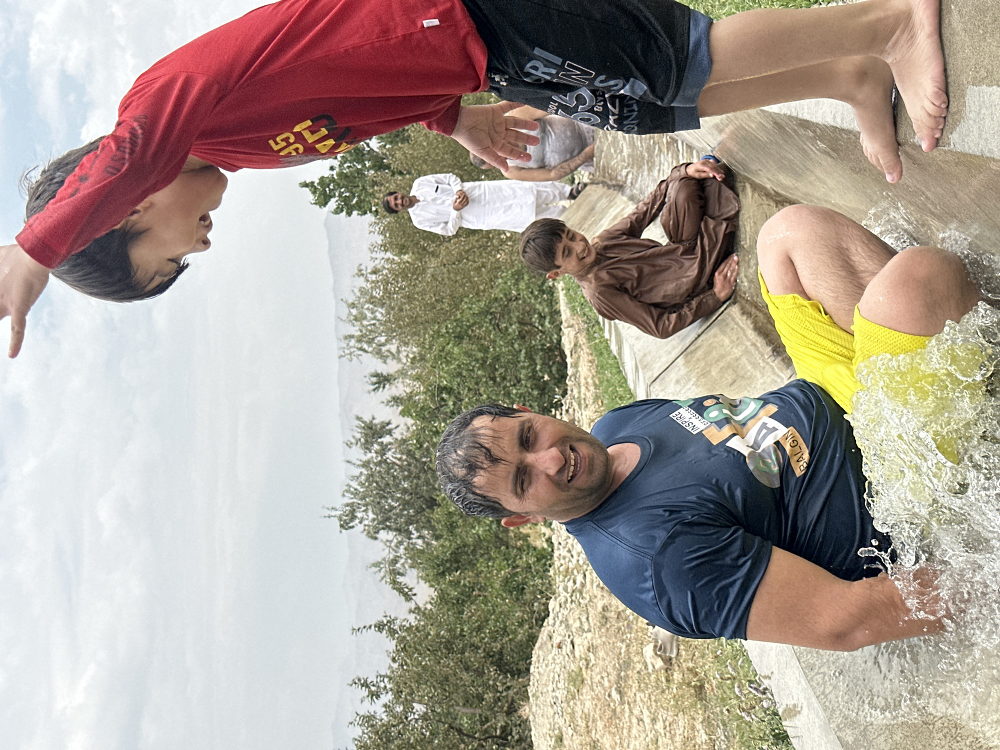 In Kan Mehtarzai, Pakistan, a group of individuals are gathered near a water channel. A man in a dark blue t-shirt and yellow shorts is partially submerged in the water, splashing it with his arm. A young boy in a brown outfit sits further down the channel, smiling. In the background, a man in a white shalwar kameez stands and smiles, while another person is partially visible in the water behind him. The environment appears to be rural, with trees and hills visible in the distance under a cloudy sky.