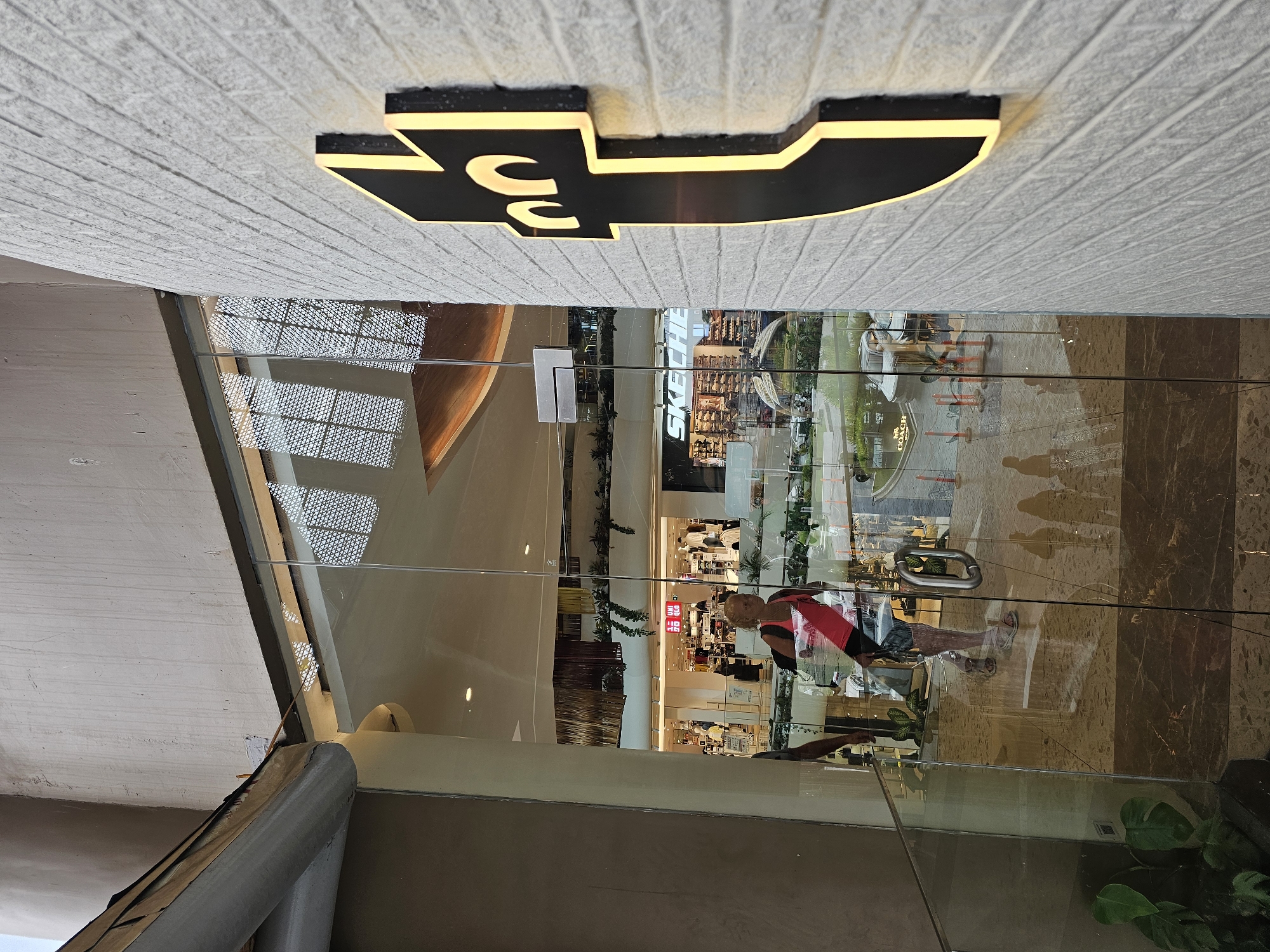 The image shows the exterior of a modern building, likely a shopping mall, in Denpasar, Indonesia. The facade features a large glass entrance with a prominent black and yellow logo mounted on a textured white brick wall. Inside the building, through the glass, several shops are visible, including a "UNIQLO" store and a "SKECHERS" store. A woman with blonde hair, wearing a pink tank top and dark shorts, is walking through the glass doors. Her reflection is visible on the glass, along with the reflections of other people and the interior of the mall. The overall impression is one of a busy commercial area in a tropical setting, suggested by the lush greenery visible in the background.