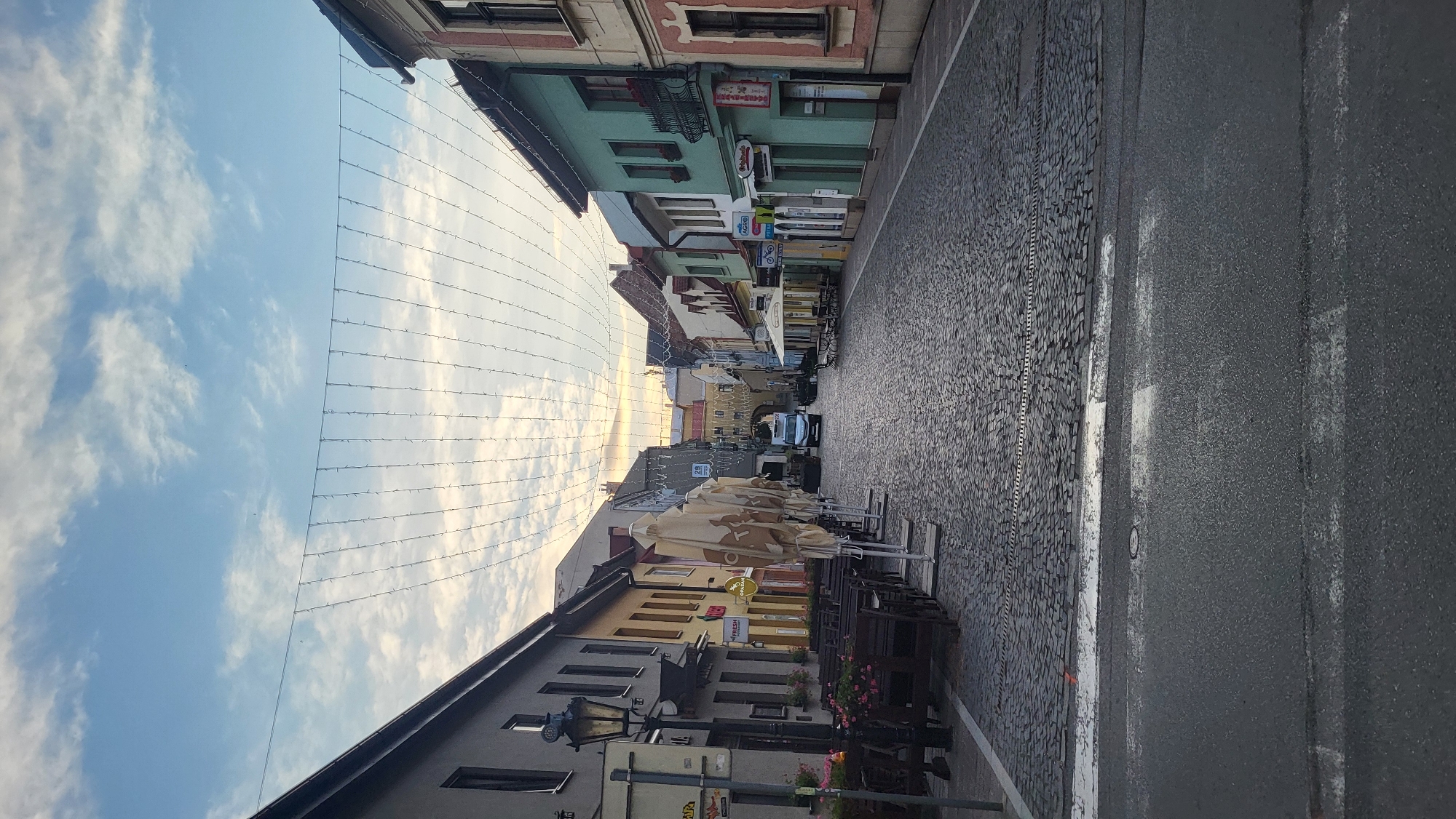 The image depicts a view down a cobblestone street in Prešov, Slovakia. The street is lined with multi-story buildings, many of which have shops or restaurants on the ground floor. String lights are strung across the street overhead, creating a festive atmosphere. Several outdoor seating areas with tables and chairs are set up along the sides of the street, some covered by umbrellas. A white van is visible driving down the street. The sky is partly cloudy, with hints of the setting sun visible in the distance. The foreground shows a paved road with pedestrian crossing markings.