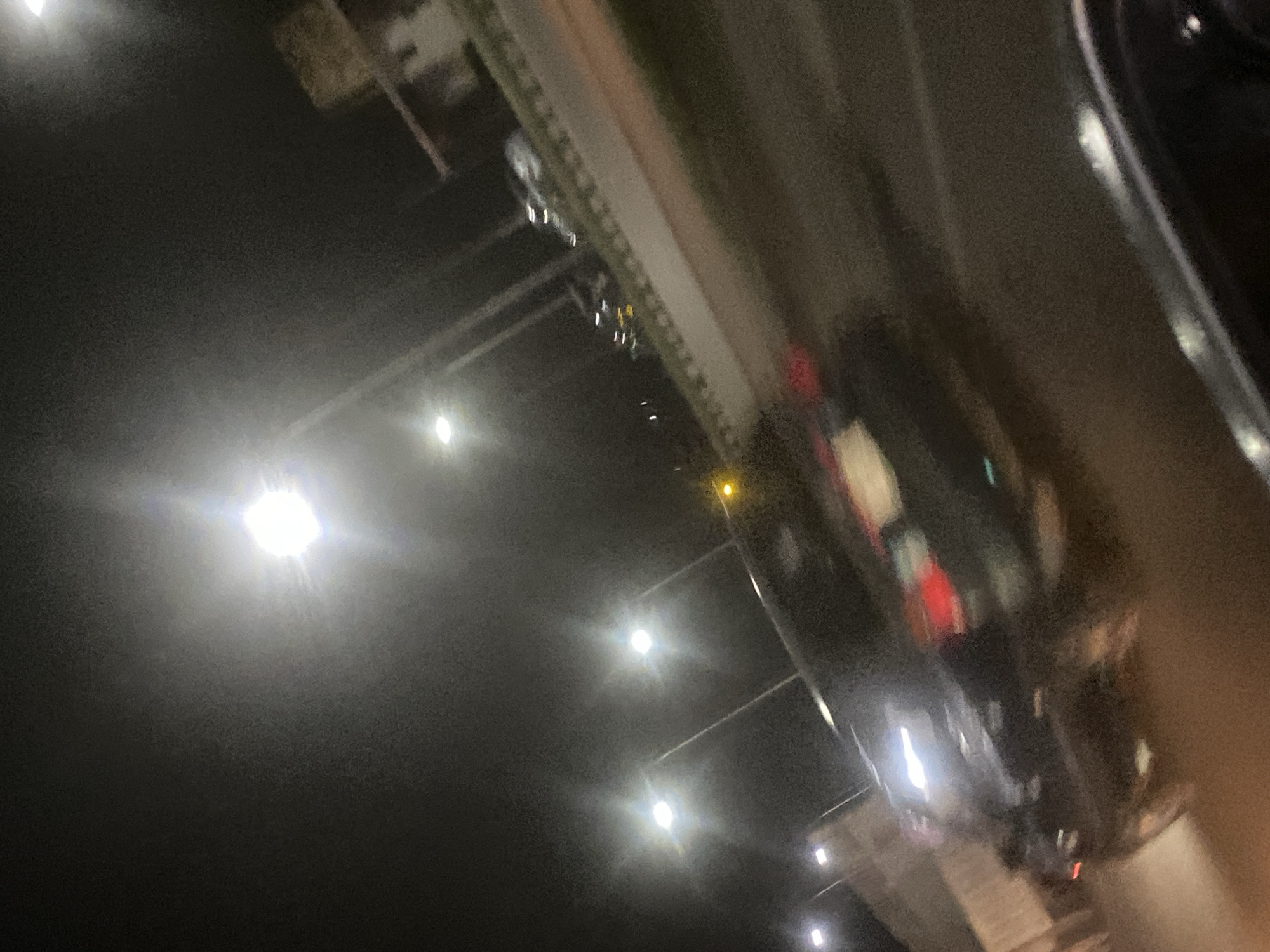 The image depicts a dark street scene in Gosa, Nigeria, at night. Streetlights illuminate the road. A line of parked cars is visible along the side of the road, with their taillights reflecting. The image is taken from a high angle and is slightly blurred, possibly from movement.