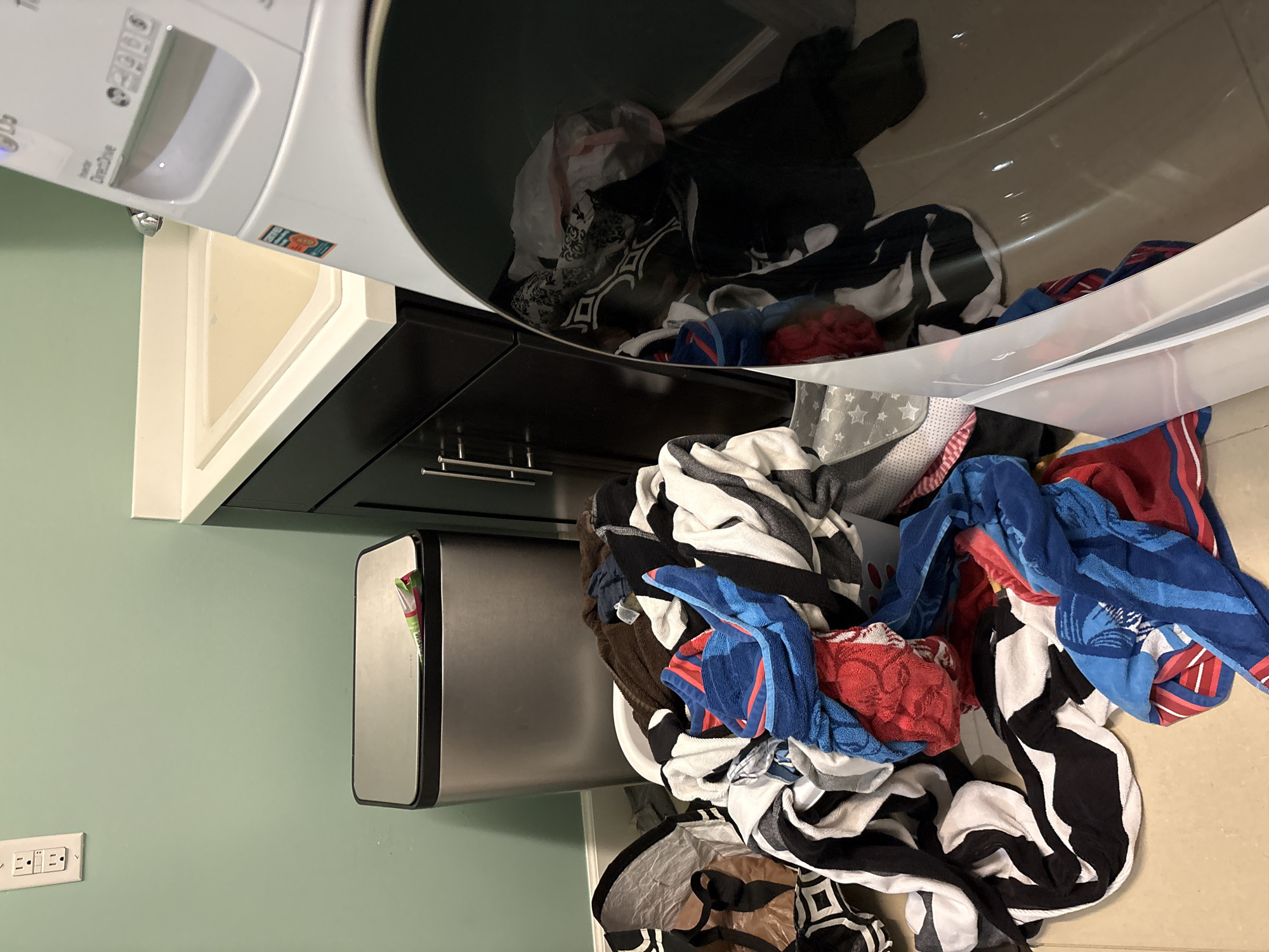 A laundry room is depicted with a washing machine in the foreground, a sink and vanity to the right, and a trash can to the left. A large pile of laundry, including towels and blankets, is spread across the floor in front of the washing machine. The room is clean and well-lit, with a pale green wall and tiled floor. This image was taken in Bradenton, Florida, United States.