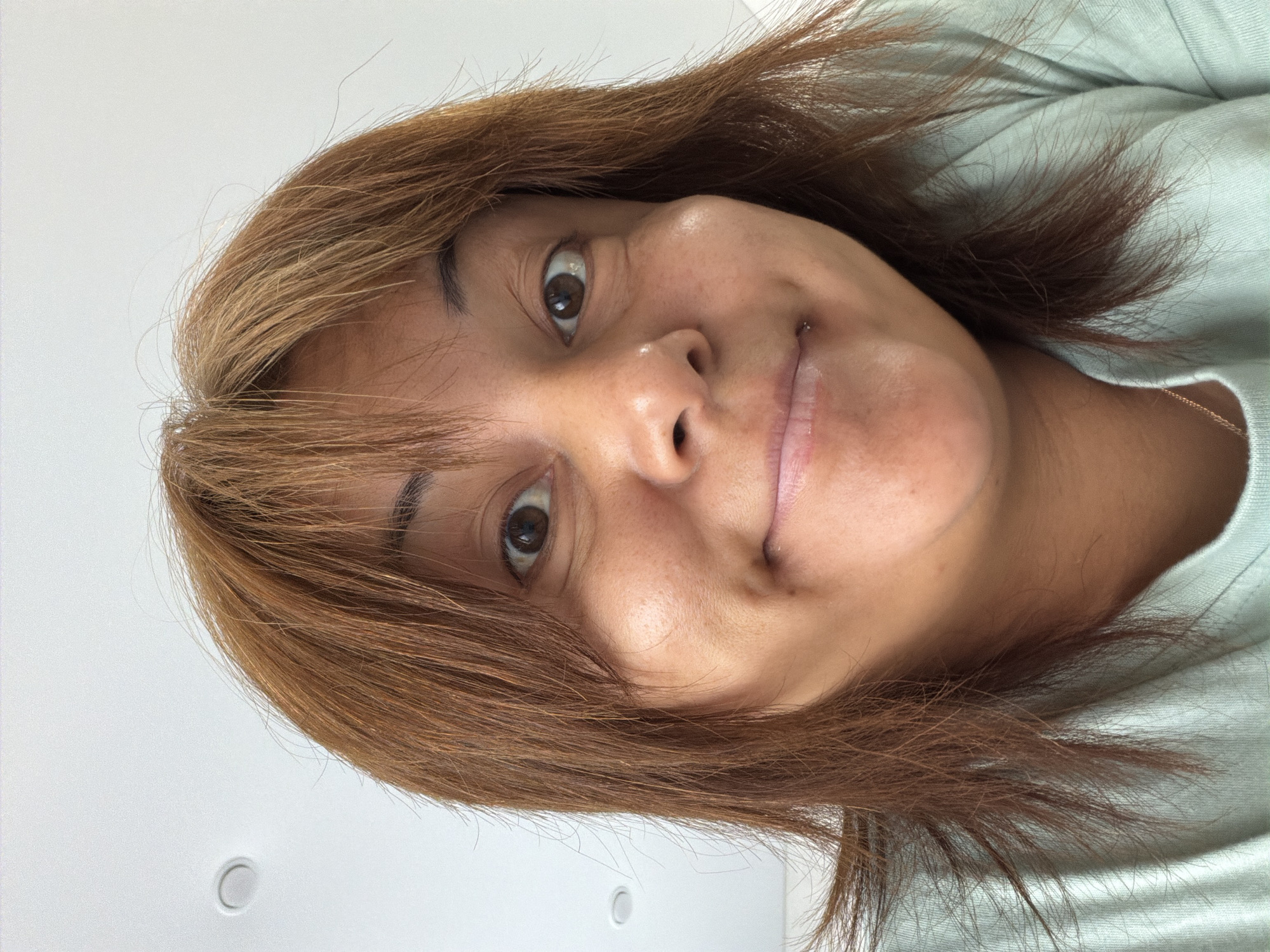 A close-up selfie of a woman with medium-length, reddish-brown hair and bangs. She has brown eyes and is smiling with her lips pursed. She is wearing a light blue t-shirt. The background is a plain white ceiling with a circular light fixture.