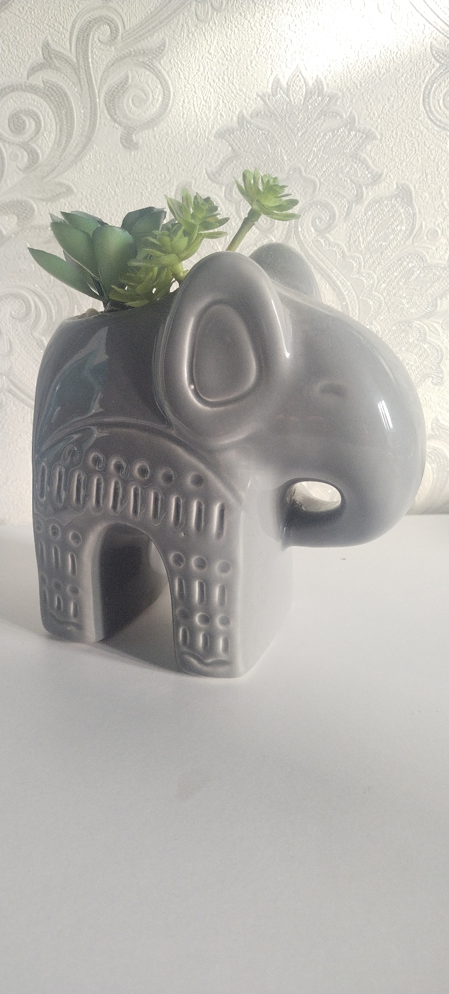 The image shows a close-up of a grey ceramic elephant-shaped planter with a succulent plant inside. The planter is positioned on a white surface, possibly a table or shelf. The background features a textured white wallpaper with a damask-like pattern. The lighting is soft and appears to be coming from the right side of the image. There are no people or any other objects visible. The scene is indoors. No information about Pisochyn, Ukraine is depicted or implied in the image.