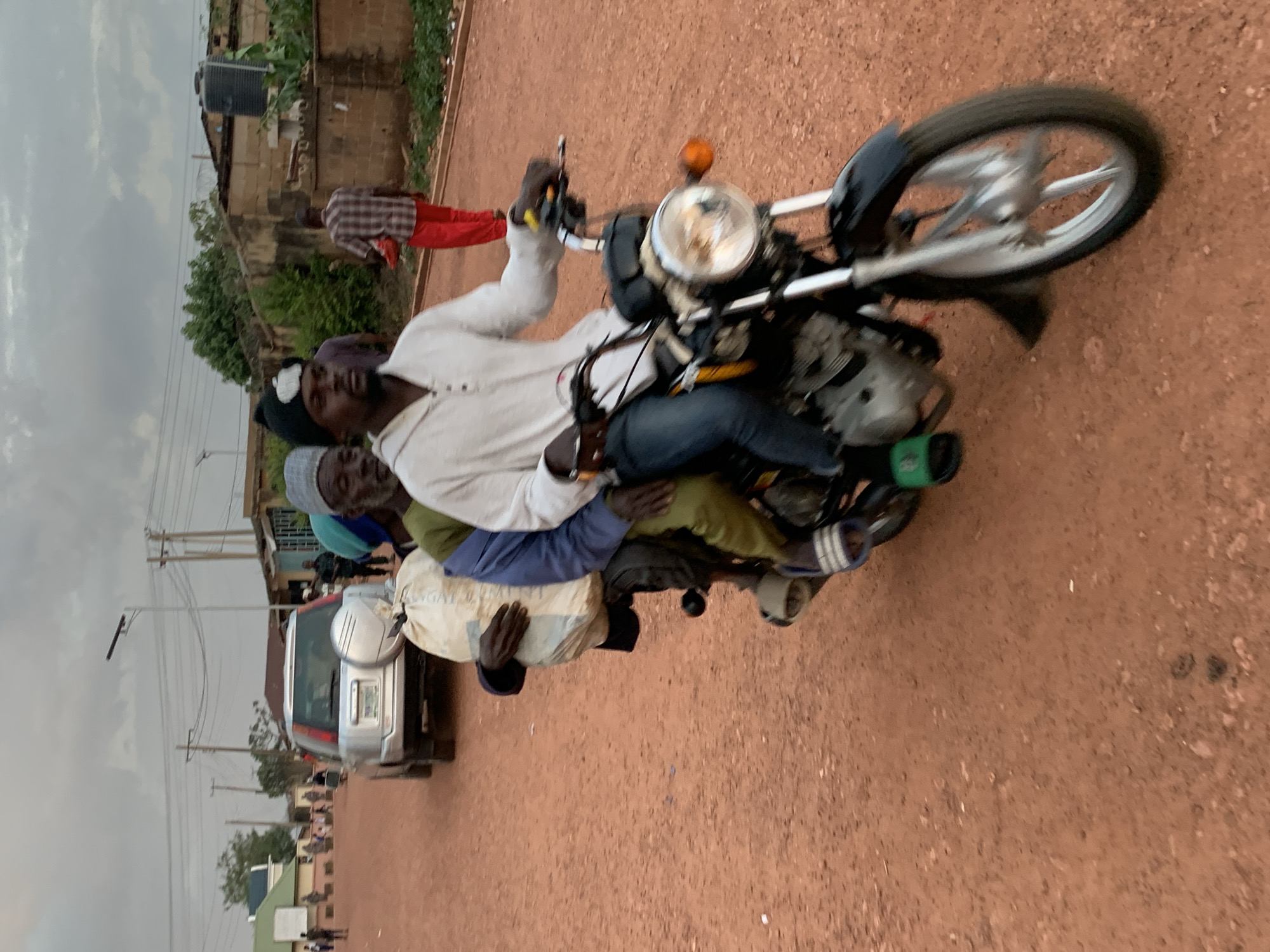 The image depicts a street scene in Gura Top, Nigeria, with a group of people riding on a motorcycle. Four individuals are visibly on the motorcycle, with the driver in front, followed by three passengers. The driver is wearing a white shirt and dark pants, while the passengers are dressed in various colors and clothing. One passenger in the middle appears to be carrying a large, light-colored bag.
The setting is an unpaved, reddish-brown road, typical of many rural or semi-urban areas in Nigeria. In the background, there are buildings constructed from brick and concrete, some with corrugated iron roofs. Trees and vegetation are also visible, suggesting a warm climate. Power lines and utility poles line the street, indicating some level of infrastructure. A silver SUV is parked on the side of the road.
The sky is overcast, hinting at a possibly cloudy or rainy day. The image is taken from a low angle, looking slightly upwards towards the motorcycle and its occupants. The overall impression is one of everyday life and transportation in a Nigerian community. There is no visible text in the image. The activity is the transportation of multiple individuals on a single motorcycle.