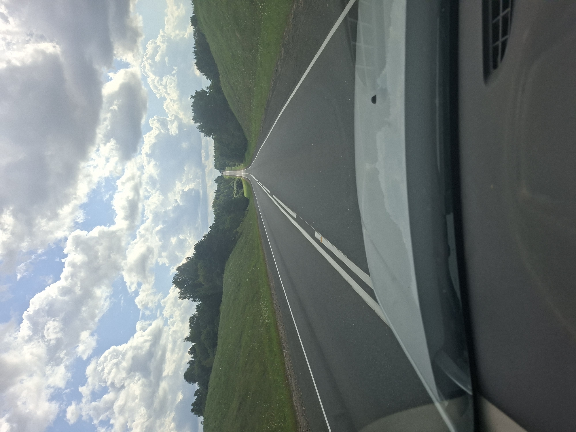 The image depicts a view from inside a vehicle, looking out through the windshield at a straight, two-lane asphalt road stretching towards the horizon. The road is lined with white dashed lines on the left and solid white lines on the right. The surrounding landscape is characterized by gently rolling green hills covered in grass, with a line of trees bordering the road on both sides, creating a natural tunnel effect in the distance. The sky above is a mix of blue with large, fluffy white clouds. The weather appears to be bright and sunny. The foreground shows the dashboard of the vehicle, a dark gray surface with a slight reflection of the sky on the windshield. The location is identified as Каменналаўскі сельскі Савет, Belarus.
