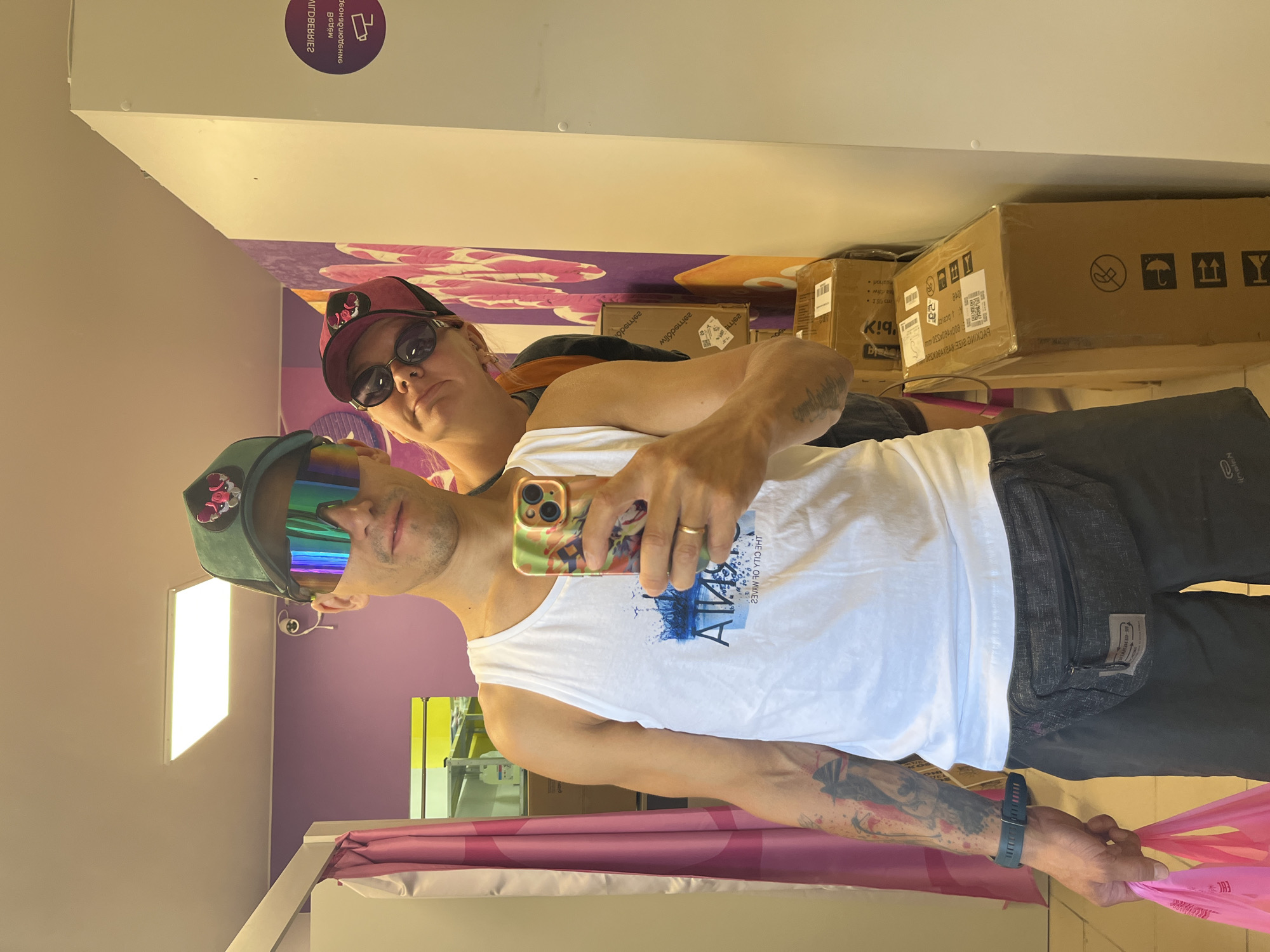 In an indoor fitting room, two individuals are captured in a mirror selfie. The person in the foreground, a male with short dark hair, wears a white tank top, dark shorts, a dark fanny pack around his waist, a dark fitness tracker on his left wrist, a green baseball cap with a logo, and futuristic-looking mirrored sunglasses. He is holding a pink plastic shopping bag in his left hand and his smartphone in his right, capturing the reflection. Behind him and slightly to the right, another person, also wearing a cap and sunglasses, leans into the frame, with only their face and upper torso visible. The fitting room features purple walls adorned with a pink floral graphic on the right, and a white partition or closet on the far right. Pink curtains hang on the left, and several cardboard boxes are stacked against the wall in the background. The lighting appears to be from overhead fluorescent fixtures. This scene does not have any specific identifying markers that would place it within the city of Chaikovsky, Russia.