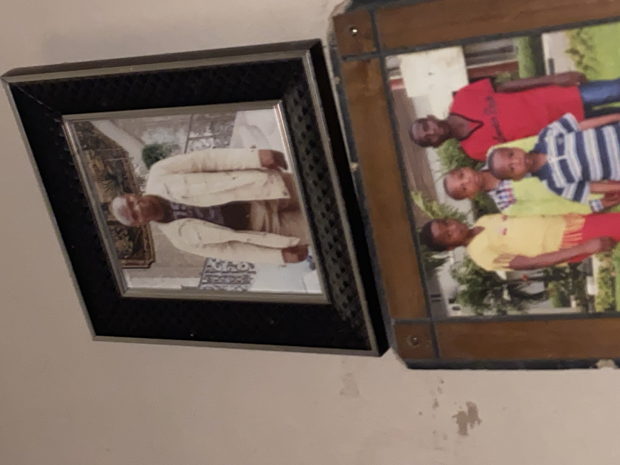 Two framed photographs are displayed on a wall. The top photograph is a portrait of a man wearing a cream-colored jacket over a dark t-shirt. He is standing and looking towards the viewer. The bottom photograph shows a group of four young males. From left to right, one boy wears a yellow shirt with fringed sleeves and red pants. Next to him, a boy in a light green shirt with a checkered pattern stands. Another boy in a red t-shirt is to the right of him, and a fourth boy in a blue and white striped shirt stands in front of the other three. All four boys appear to be looking towards the camera. The background of the bottom photograph shows a building and greenery. The wall appears to be painted a light color and shows some signs of wear. The location is identified as Bukuru, Nigeria.
