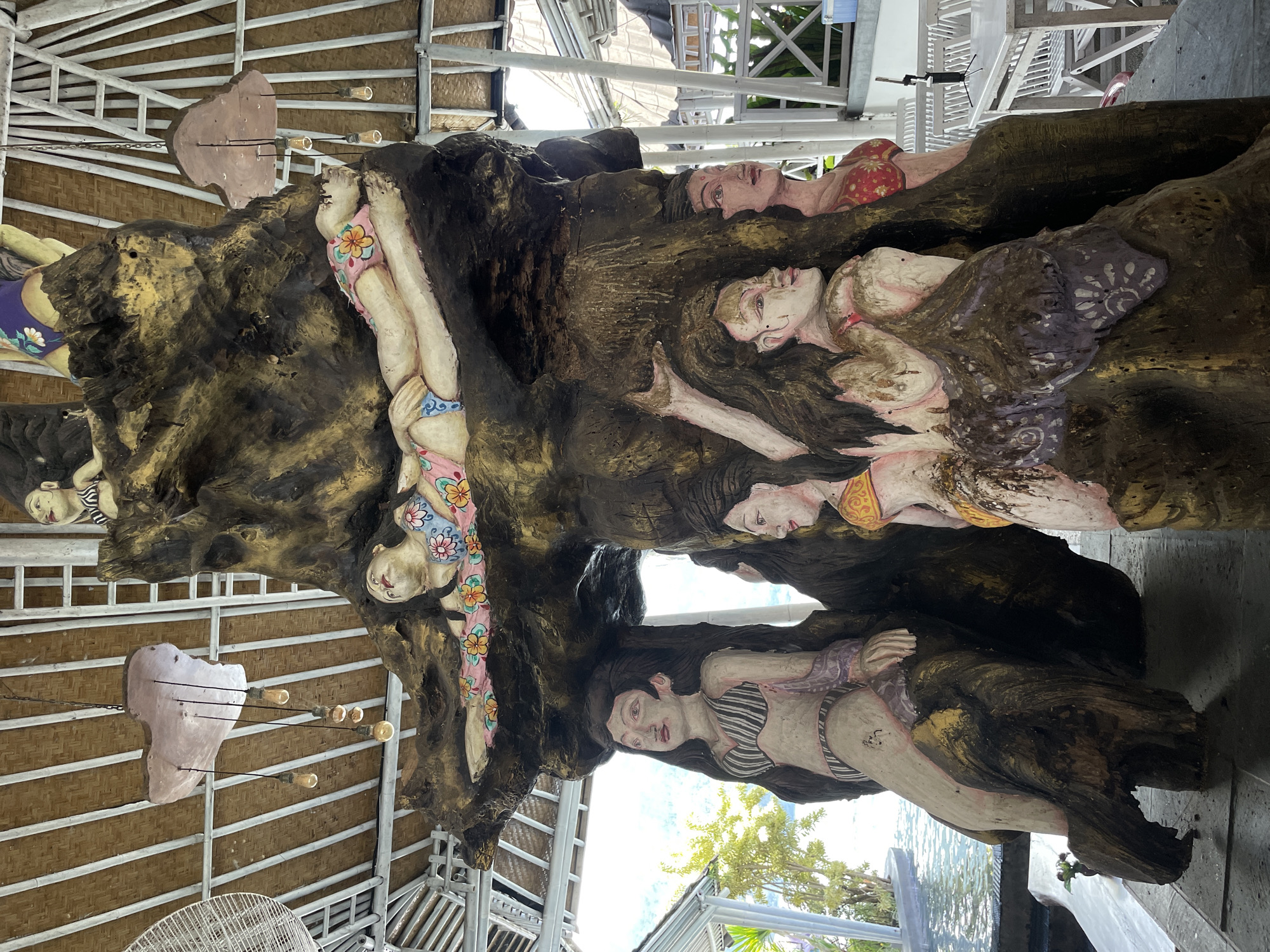 The image depicts a large, ornately carved wooden structure featuring several human figures, seemingly women in various states of repose and attire, carved into the wood. The figures are painted with bright colors, particularly in their swimwear. The structure appears to be located in an open-air building with a bamboo and metal frame roof. The setting suggests a tropical or coastal environment. The location of the city of Kintamani, Indonesia, is mentioned.