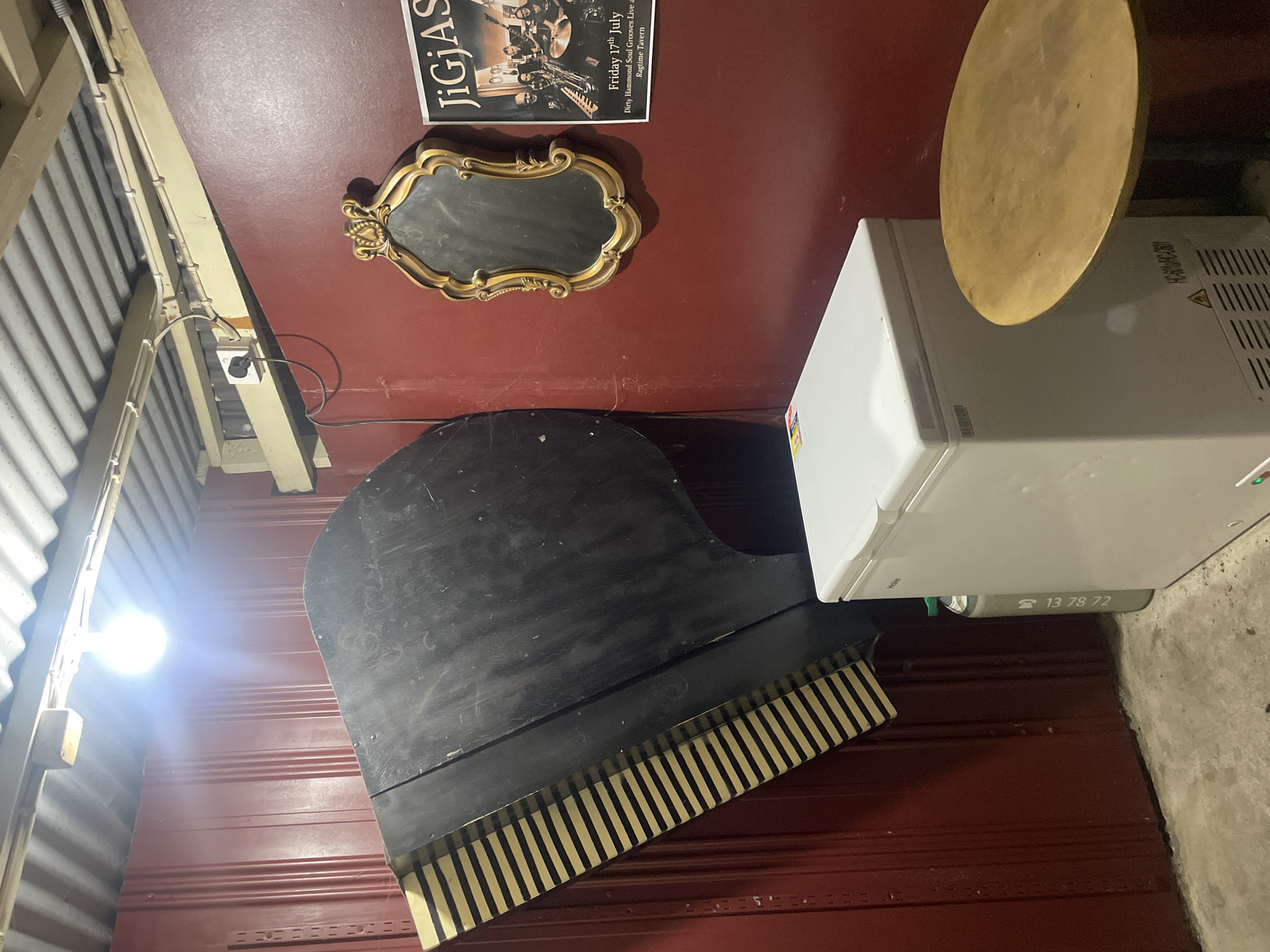 The image depicts the interior of a dimly lit establishment, possibly a bar or music venue, in Melbourne, Australia. A large, black grand piano lid is propped at an extreme angle against a dark red corrugated metal wall, with its keys facing upwards and outwards. To the right of the piano lid, a white chest freezer stands on a concrete floor. Leaning against the freezer is a tall, narrow green cylinder with white lettering. Above the freezer, a framed, ornate gold mirror hangs on the red wall. To the right of the mirror is a poster advertising a musical event titled "JiGjAS" on "Friday 17th July" at "Dirty Hammond Soul Grooves Live at Ragtime Tavern". A small, round, light brown wooden table with a metal base is positioned to the right of the freezer. The ceiling is corrugated metal, with exposed wiring and a bright, bare light bulb illuminating the scene from the top left. The overall impression is one of a somewhat cluttered and unconventional storage or preparation area for a performance space.