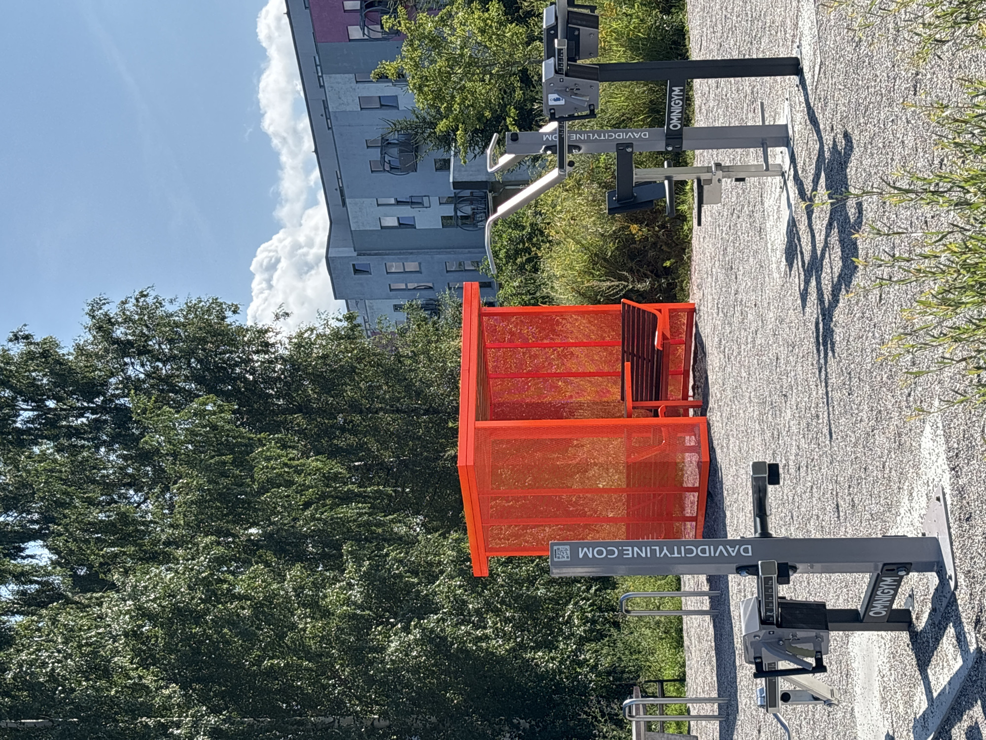 This is an outdoor scene at a fitness park in Tallinn, Estonia. In the foreground, there are several pieces of outdoor gym equipment, including a leg press machine and a chest press machine, both labeled "OMNIGYM". The ground is covered in small gravel. To the left, there are bicycle racks. In the mid-ground, there is a bright orange, open-sided shelter with a bench inside. A vertical sign next to the shelter displays "DAVIDCITYLINE.COM" and a QR code. In the background, there is lush green foliage, tall trees, and a modern, multi-story apartment building with balconies. The sky is blue with some scattered white clouds.
