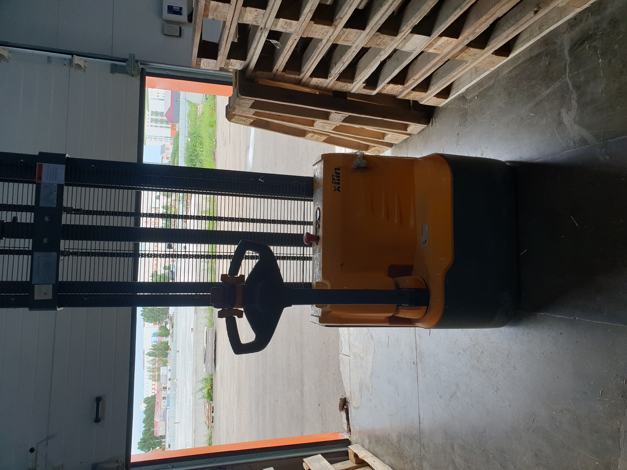 The image shows an industrial setting, likely a warehouse or loading dock, with a yellow and black electric pallet jack in the foreground. The pallet jack is positioned centrally, facing towards an open garage door that leads to an outdoor area. To the right of the pallet jack, a stack of wooden pallets is visible, leaning against a wall. The floor is concrete, with some debris scattered around. Outside the open door, a paved area with buildings and trees in the background can be seen. The scene appears to be an overcast day. There are no people present in the image. The location is identified as Aktobe, Kazakhstan.