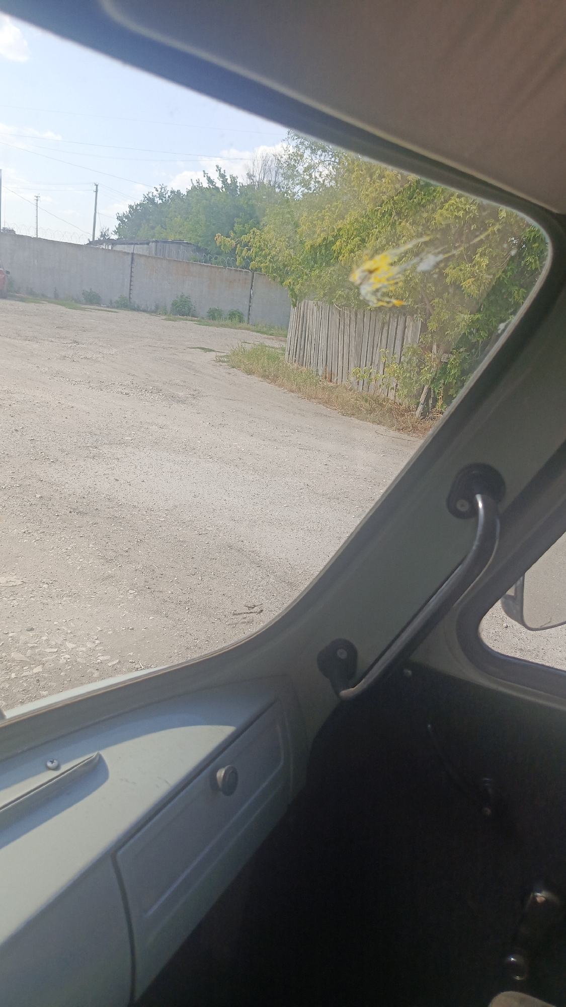 The image is taken from inside a vehicle, looking out through the windshield. The scene outside is an unpaved road with a concrete wall and a wooden fence on either side. Trees and vegetation are visible behind the fence. There is a bird dropping on the windshield. The interior of the vehicle shows a dashboard and a door handle. The lighting suggests it is daytime. The location is the city of Zolotaya Step, Russia.