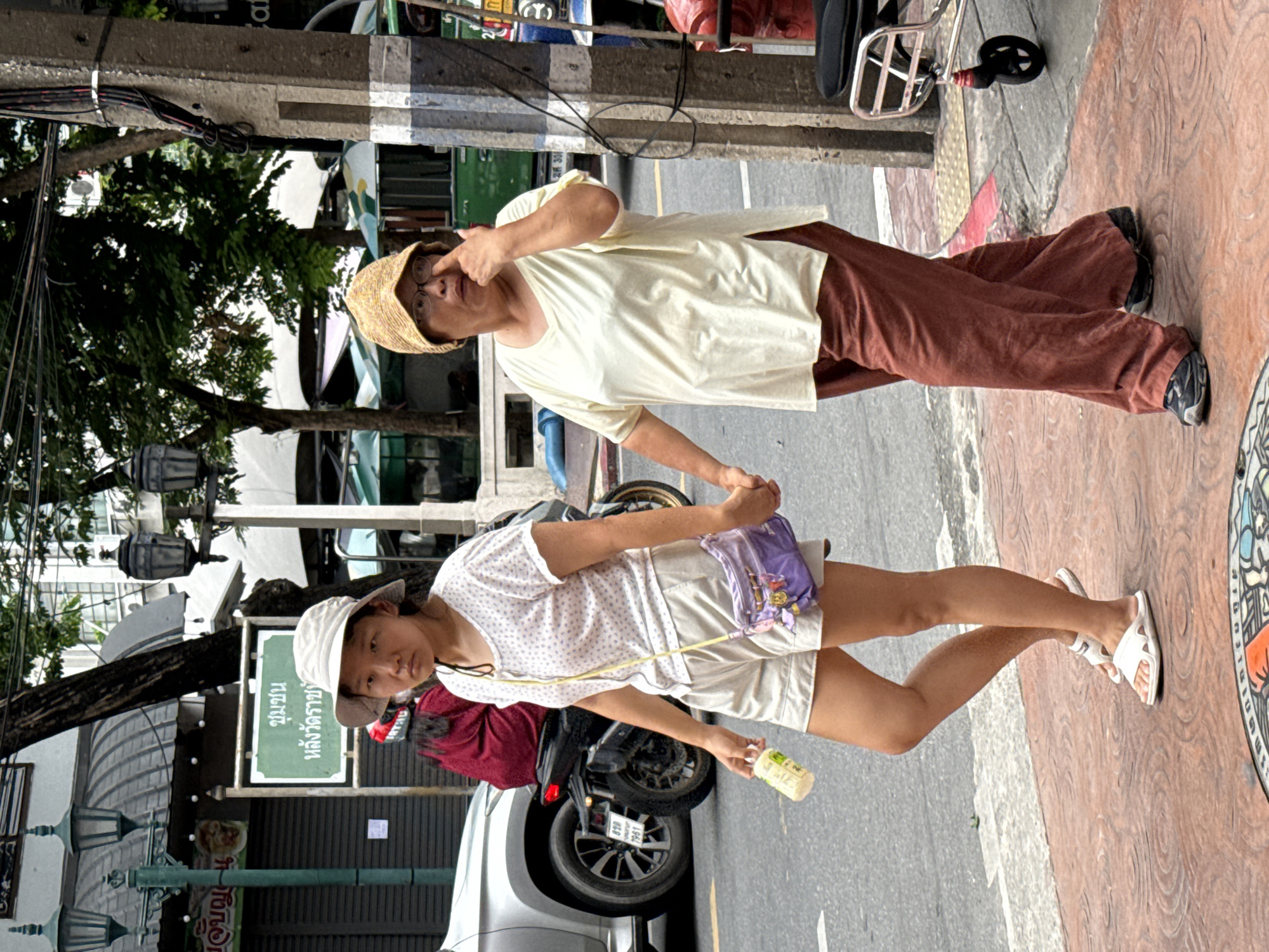 Two women, one younger and one older, walk side-by-side on a sidewalk in Bangkok, Thailand. The younger woman, on the left, wears a white patterned t-shirt, beige shorts, and white flip-flops. She carries a light-colored bottle in her right hand and a small purple purse hangs from a yellow strap across her body. The older woman, on the right, wears a cream-colored t-shirt, rust-colored wide-legged pants, and dark athletic shoes. She has a straw hat on her head and her right hand is raised to her face, touching her eye. They are holding hands. In the background, a silver car and a motorcycle are visible on the street. The sidewalk is paved with reddish-brown tiles with a circular pattern. A concrete utility pole with wires is on the right side of the image. Signs in Thai are visible on a building in the background, including one with green lettering on a white background. Trees and awnings are also part of the urban environment.