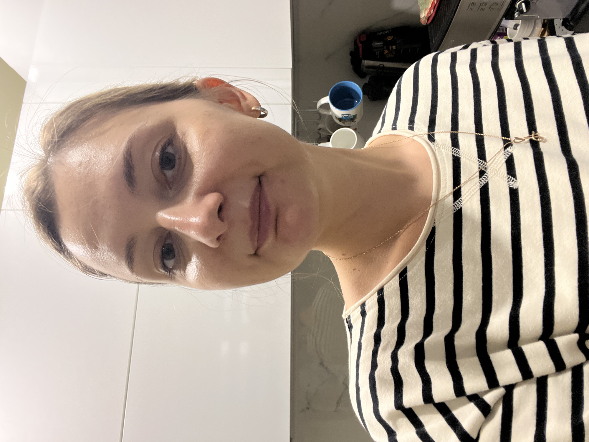 The media file is a portrait of a young woman, facing slightly away from the camera and looking to her right. She has light brown hair pulled back and is wearing a black and white striped top. A delicate gold necklace with a small charm is visible around her neck. Her expression appears neutral or perhaps slightly introspective.
In the background, there are hints of a kitchen or bathroom setting. On a shelf or counter, a blue and white mug and a white cup are visible. A black appliance, possibly a coffee maker or a kitchen mixer, is also present. The environment appears to be indoors, with the lighting suggesting it is daytime. The surfaces are clean and modern, with white cabinetry and grey tiled walls. There are no strong indicators of time of day or weather, but the indoor setting implies a controlled environment. The overall mood is calm and personal. The location context of Volgograd, Russia, does not offer specific visual cues within the image itself. There is no discernible text in the image.