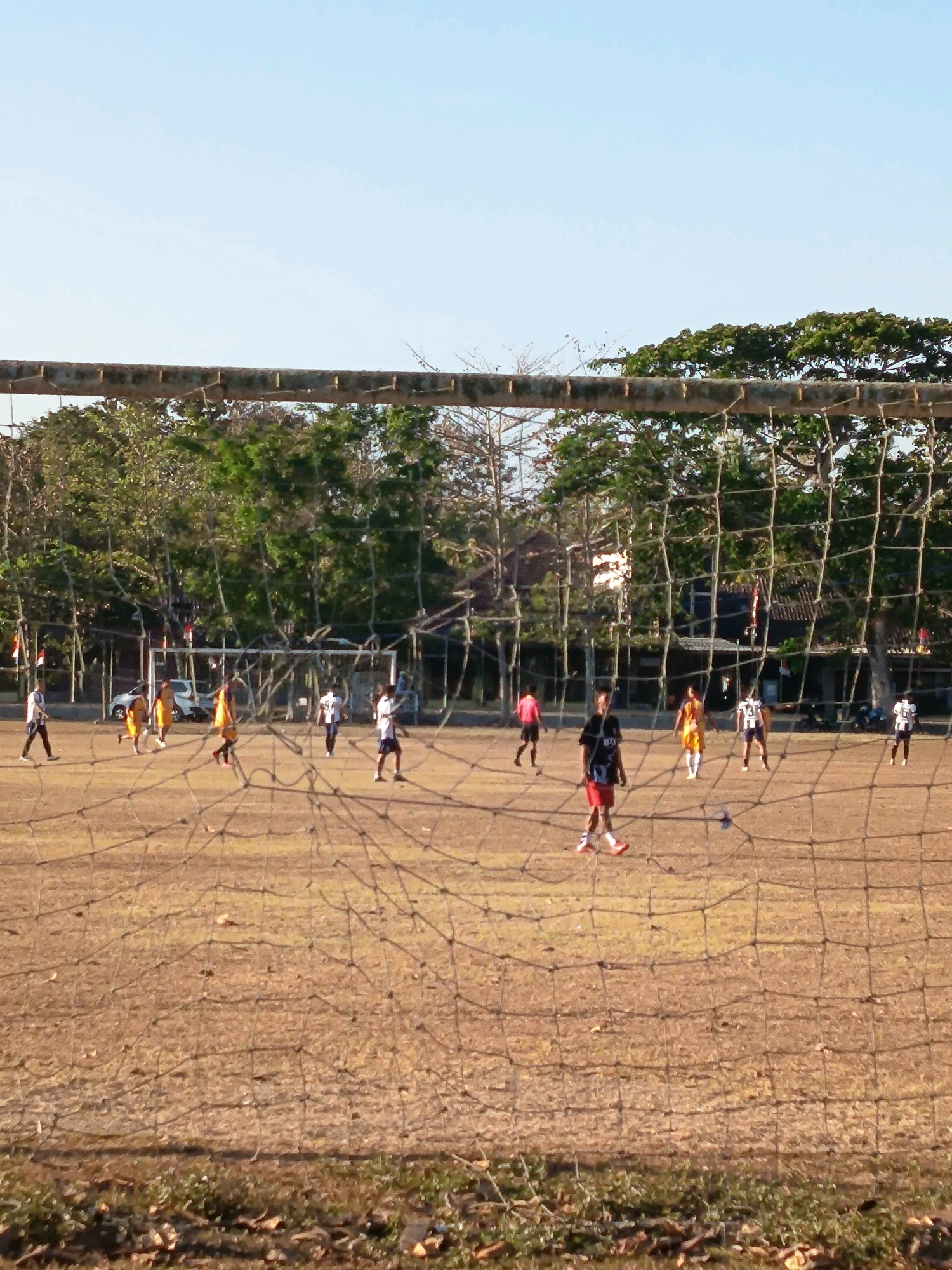 The image captures a scene of a soccer game being played on a dry, dirt field in Pengasih, Indonesia. The field is surrounded by trees and features a goalpost with a net in the foreground, suggesting this is a local or community sports area. Several young men, dressed in various soccer attire, are actively engaged in the game. Some are running, while others appear to be positioned on the field, possibly waiting for the ball or observing the play. The backdrop includes more trees and some buildings, hinting at the surrounding environment of Pengasih. The lighting suggests it is daytime, with the sun casting shadows on the field. The overall impression is one of a casual, local sporting event taking place in a natural setting.