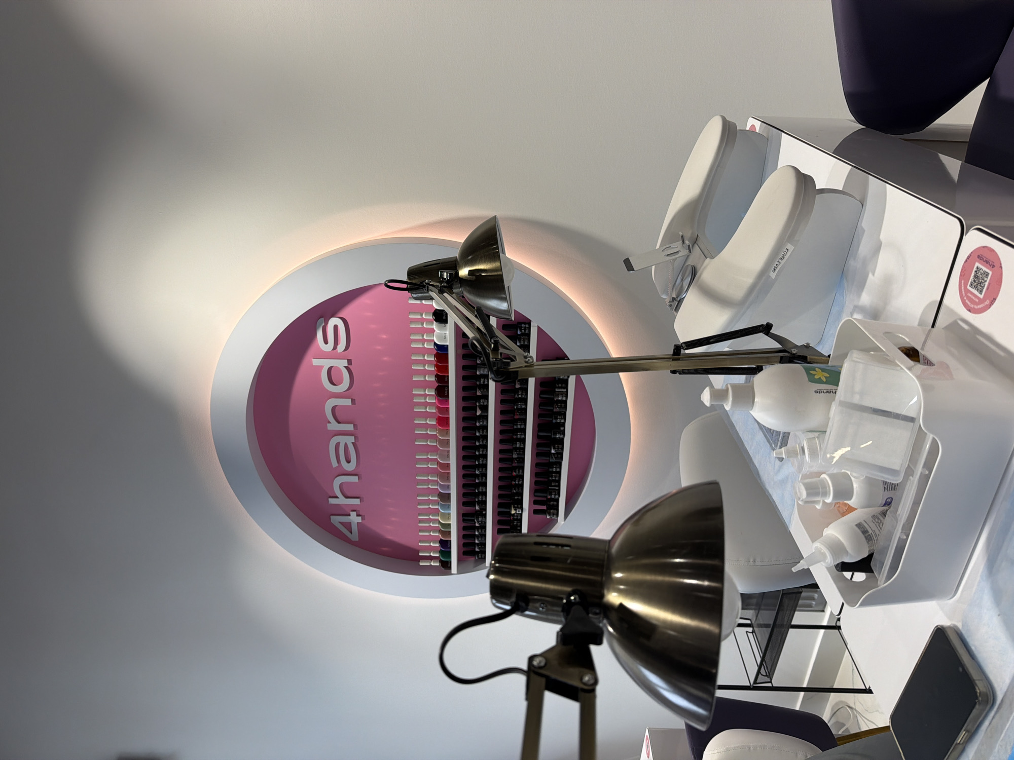 The image captures an interior view of a beauty salon or nail studio, likely located in Khimki, Russia, focusing on a manicure or pedicure station. The central feature is a large, circular, backlit sign mounted on a light-colored wall, displaying the brand name "4hands" in white, upright text against a pink background. Directly behind the sign, numerous rows of nail polish bottles, in both light and dark shades, are neatly arranged on shelves.
In the foreground and mid-ground, two metallic, adjustable desk lamps with dome-shaped shades are prominent. To the right, a white manicure/pedicure station is set up, featuring white, padded footrests or handrests. One of these supports has text partially visible, appearing to be "KOVALEVSK". A white caddy filled with various white product bottles, likely lotions or cleansers, sits on the station. A smartphone is also visible on the surface. In the upper right corner, a person's legs, clad in dark trousers or leggings, are visible, suggesting a client is seated at the station. A circular sticker featuring a QR code and the "4hands" logo is also affixed to the station. The scene depicts a clean, modern, and well-lit environment, indicating a professional setting for nail care services.
