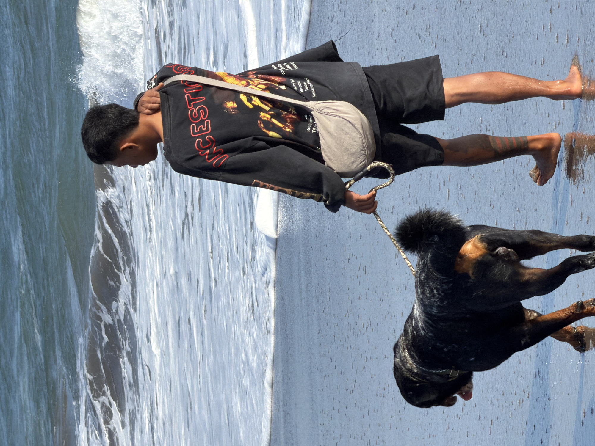 A man and his Rottweiler dog walk along the wet sand of a beach in North Kuta, Indonesia. The man, with short dark hair and tattoos on his legs, wears a black long-sleeved shirt with a graphic print on the back and black shorts. He is holding a leash attached to the dog. The Rottweiler is black with tan markings and has its tongue hanging out. The ocean behind them features waves and foam. The scene is captured from a medium distance, focusing on the man and dog from the knees up.
