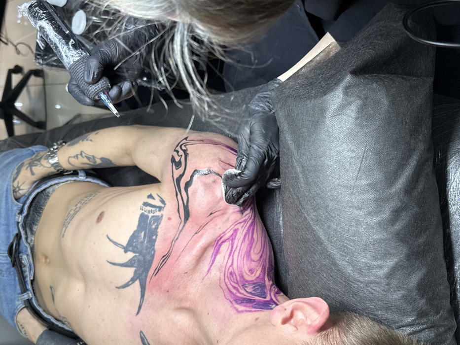 The image captures a tattoo session in progress within a studio located in Rostov-on-Don, Russia. A male subject is lying supine on a dark, fabric-covered examination table, wearing blue denim jeans. His torso is exposed, revealing existing extensive dark tattoos across his chest and arms, including a prominent abstract design on the left upper chest and multiple band-style tattoos on the right arm. A watch is present on his right wrist.
A tattoo artist, partially visible on the right side of the frame with light-colored hair, is actively working on the client's left upper chest, collarbone, and neck area. The artist wears black protective gloves. Their right hand operates a black, plastic-wrapped tattoo machine, which has its needle engaged with the client's reddened skin. The artist's left gloved hand holds a white wipe against the client's skin near the working area. A purple, swirling stencil pattern is distinctly visible on the client's lower neck and upper chest, indicating the design in progress. Black line work, consistent with the tattoo design, is already present on the upper left shoulder/collarbone. In the background, a dark five-wheeled office chair is partially visible.