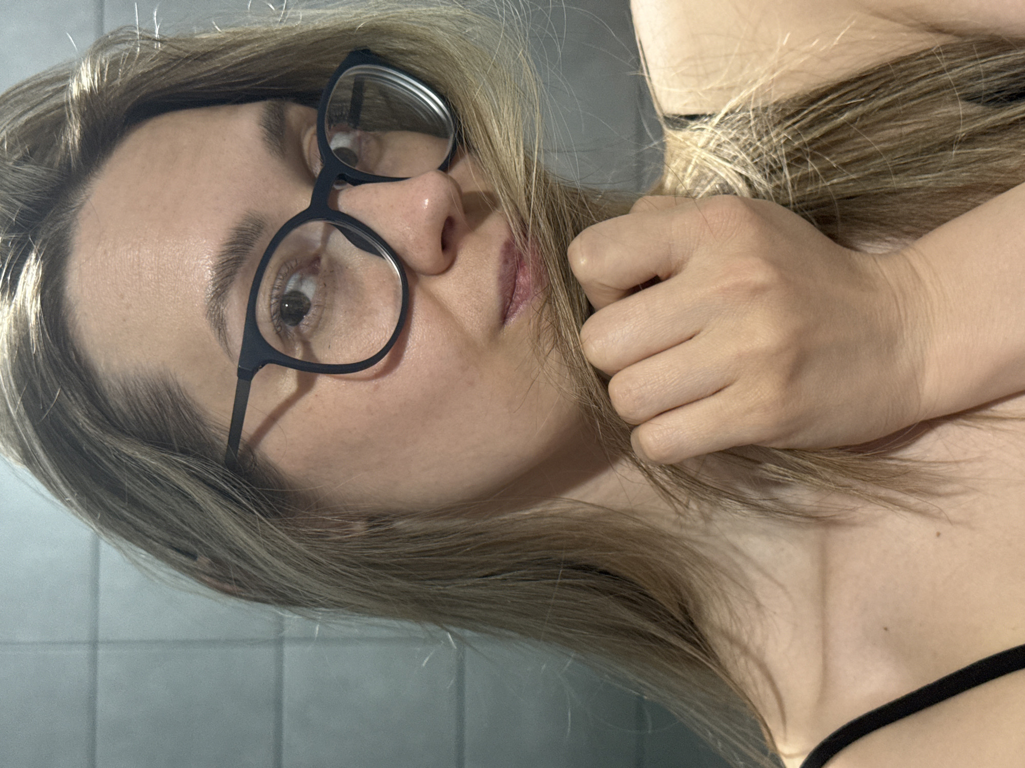 A woman with light brown hair and glasses poses for a selfie. She is wearing a black tank top. The background appears to be a gray tiled wall. The location is not identifiable as Chaikovsky, Russia, based on the visual information provided.