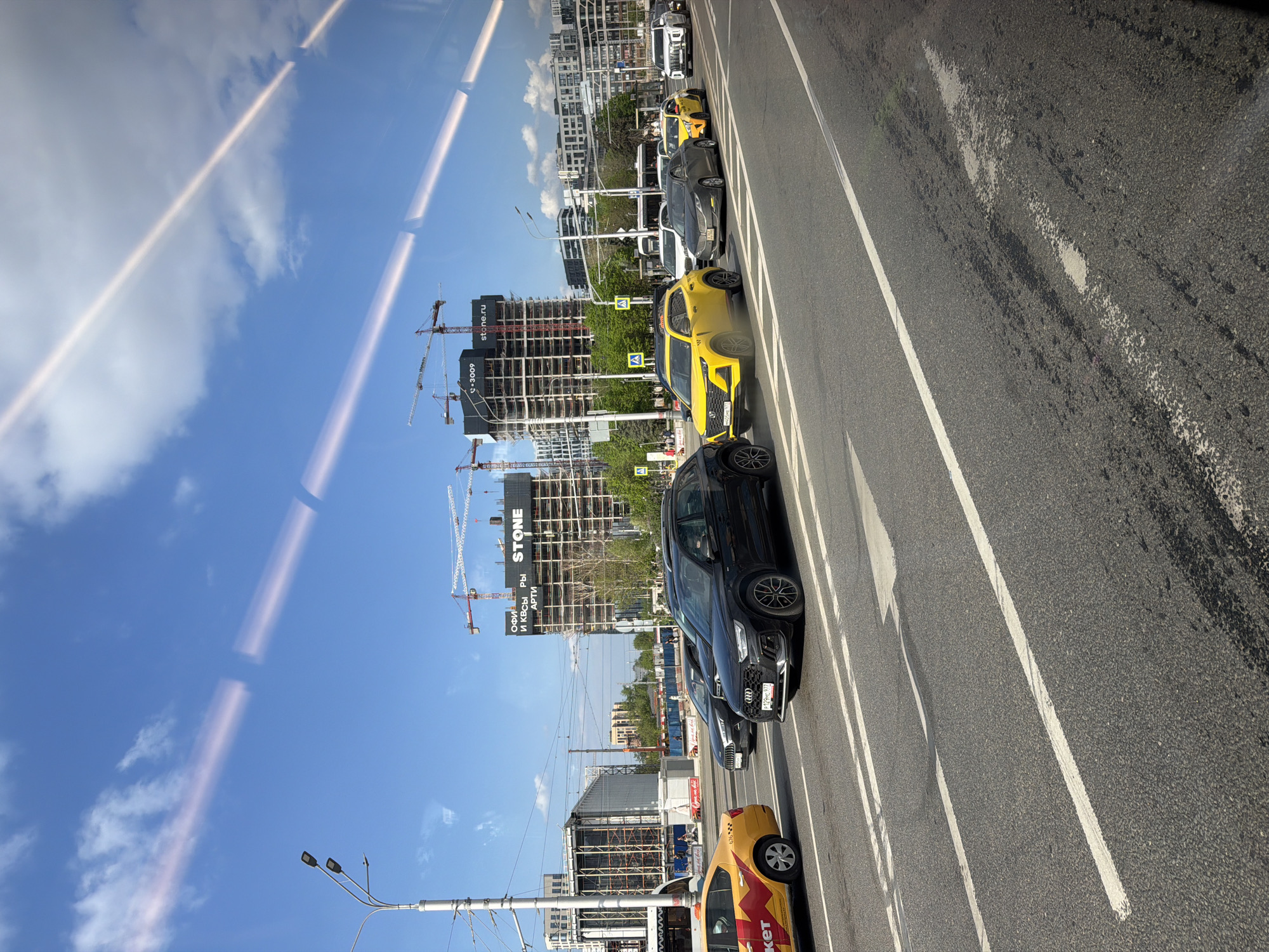 The image shows a street scene in Moscow, Russia, likely taken from a moving vehicle. A line of vehicles, including taxis, are positioned in traffic lanes marked by white lines on asphalt. A construction site is visible in the background, featuring a building under construction with several cranes. A large sign with the word "STONE" is attached to the building. Streetlights are also visible. The sky is blue with some cloud cover. The image appears to be a reflection in a window or glass surface, as indicated by the vertical lines and reflections of the sky.
