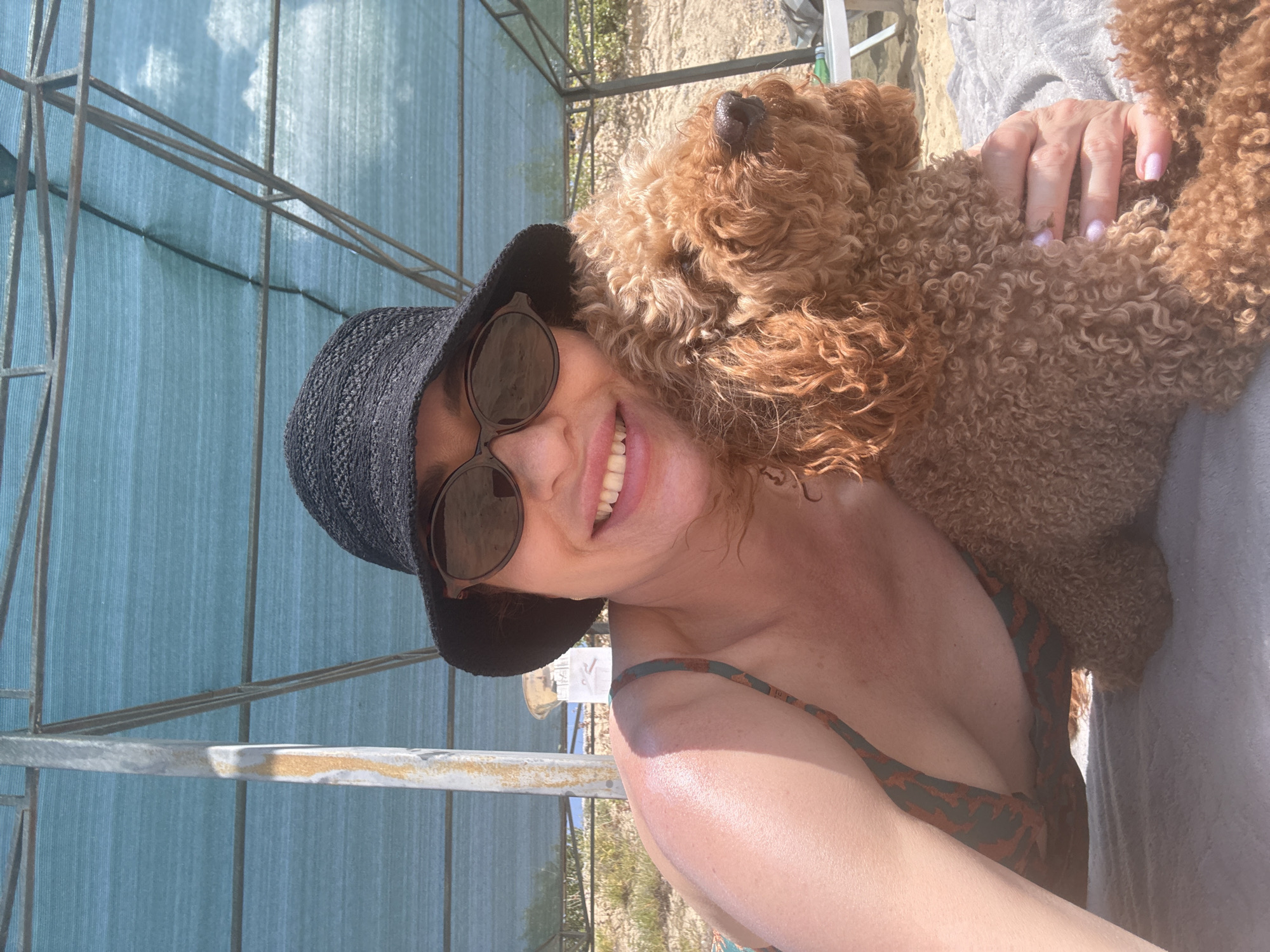 A woman with curly red hair, wearing a black hat and sunglasses, smiles at the camera while embracing a brown poodle. She is wearing a patterned swimsuit. They are lying on a gray blanket under a blue canopy structure. The background shows a sandy beach with some vegetation and a beach chair. The scene appears to be a sunny day.