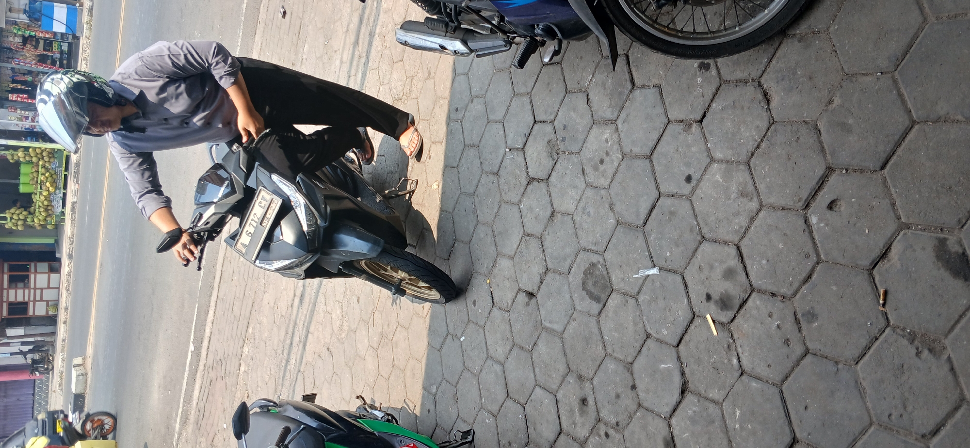 A person wearing a grey collared shirt, black pants, and a silver helmet is standing next to a dark-colored scooter on a sidewalk. The scooter has a license plate that reads "A 6 712 GY". The sidewalk is paved with hexagonal concrete tiles. In the background, there is a street with vehicles and shops, including one with a display of durians. There are also other scooters parked on the sidewalk. The scene appears to be in a city in Indonesia, possibly Baros, given the license plate format.