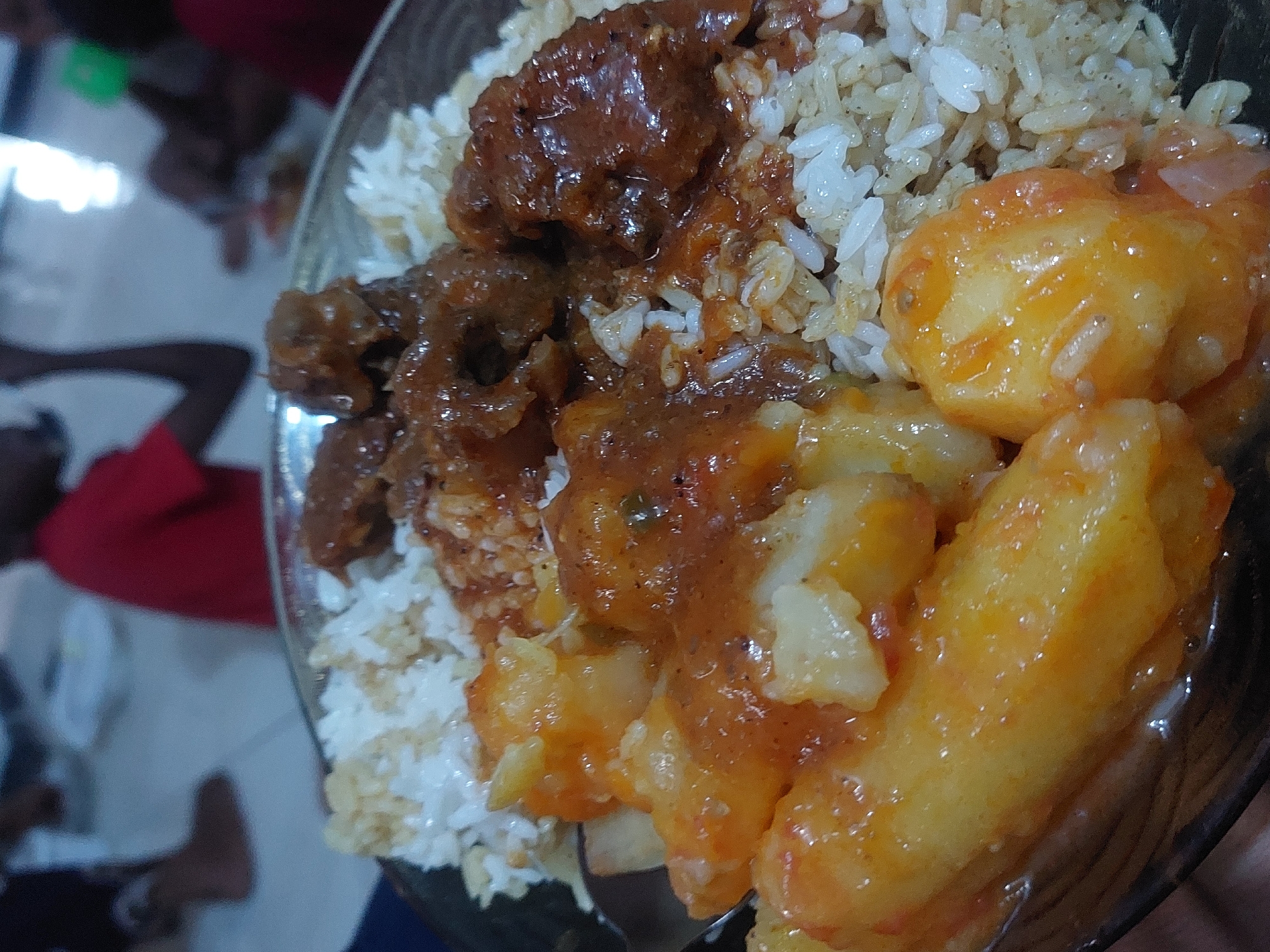 Here is a detailed description of the image:
**Scene:** A close-up shot of a plate of food, likely taken indoors, possibly in a home setting. Unidentifiable persons are in the background.
**Food Items:** The plate contains rice, what appears to be stewed meat (possibly beef), and cooked potatoes in a sauce.
**Location Clues:** While the image doesn't provide explicit location details, if the location is Dar es Salaam, Tanzania, the dish could be a common meal found in the region, such as rice with meat stew and potatoes in a tomato-based sauce.
**Objects:** Besides the food, a plate.
**People:** There are partially visible individuals in the background.