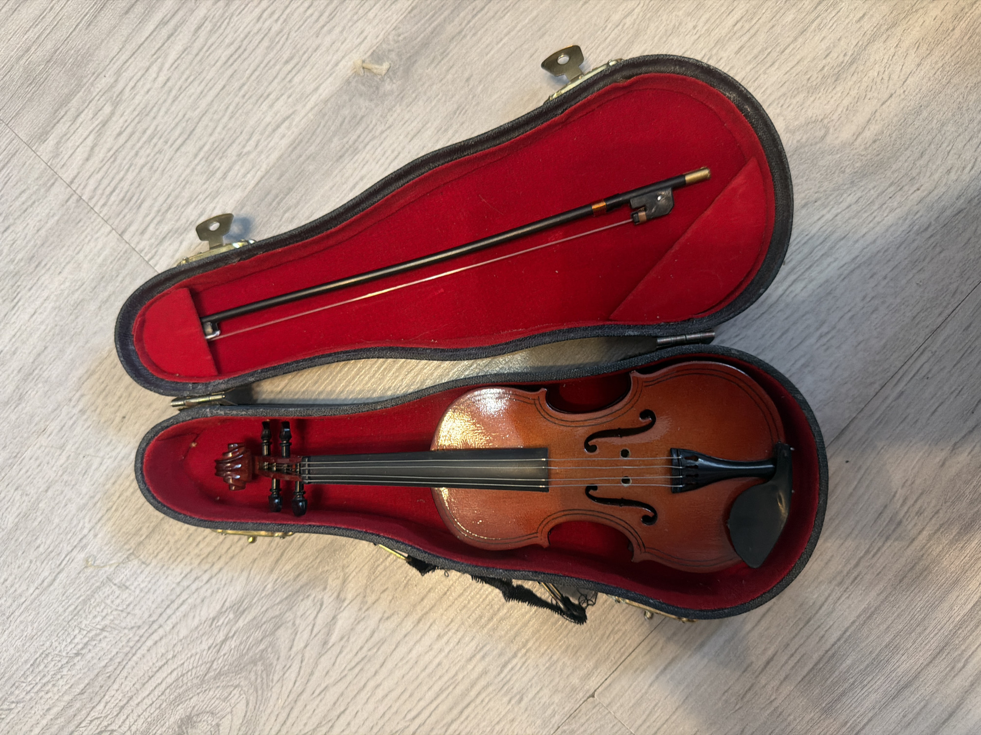 An overhead shot shows a small, brown violin and a black bow resting inside an open, red-lined violin case. The case is dark gray on the outside and appears to be made of a faux leather material. It is positioned on a light-colored wooden floor. The violin is placed in the left half of the case, with its scroll towards the top and its body extending downwards. The bow is laid out in the right half of the case, with the frog (the part held by the hand) towards the bottom. The background is the wooden floor.