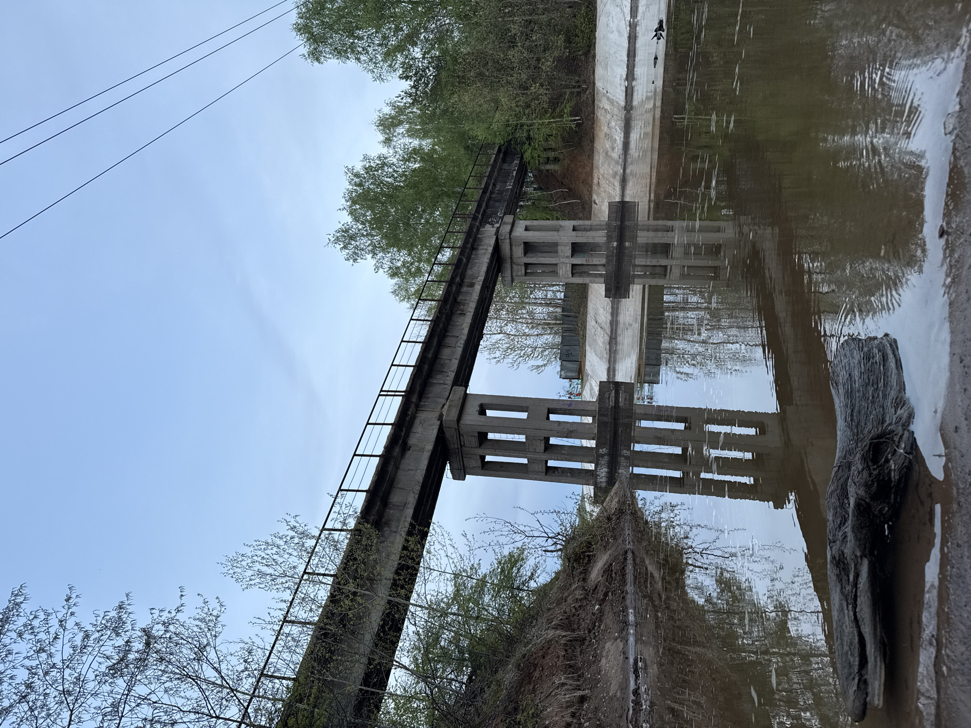 A landscape photograph of an abandoned concrete aqueduct bridge in an unknown city. The bridge spans over a body of water that reflects the structure. The bridge is supported by concrete columns with rectangular openings. A metal railing runs along one side of the bridge. Trees and vegetation surround the bridge on both sides. The sky is overcast. Power lines are visible in the upper left corner of the image. A piece of driftwood is present in the lower right corner of the frame.