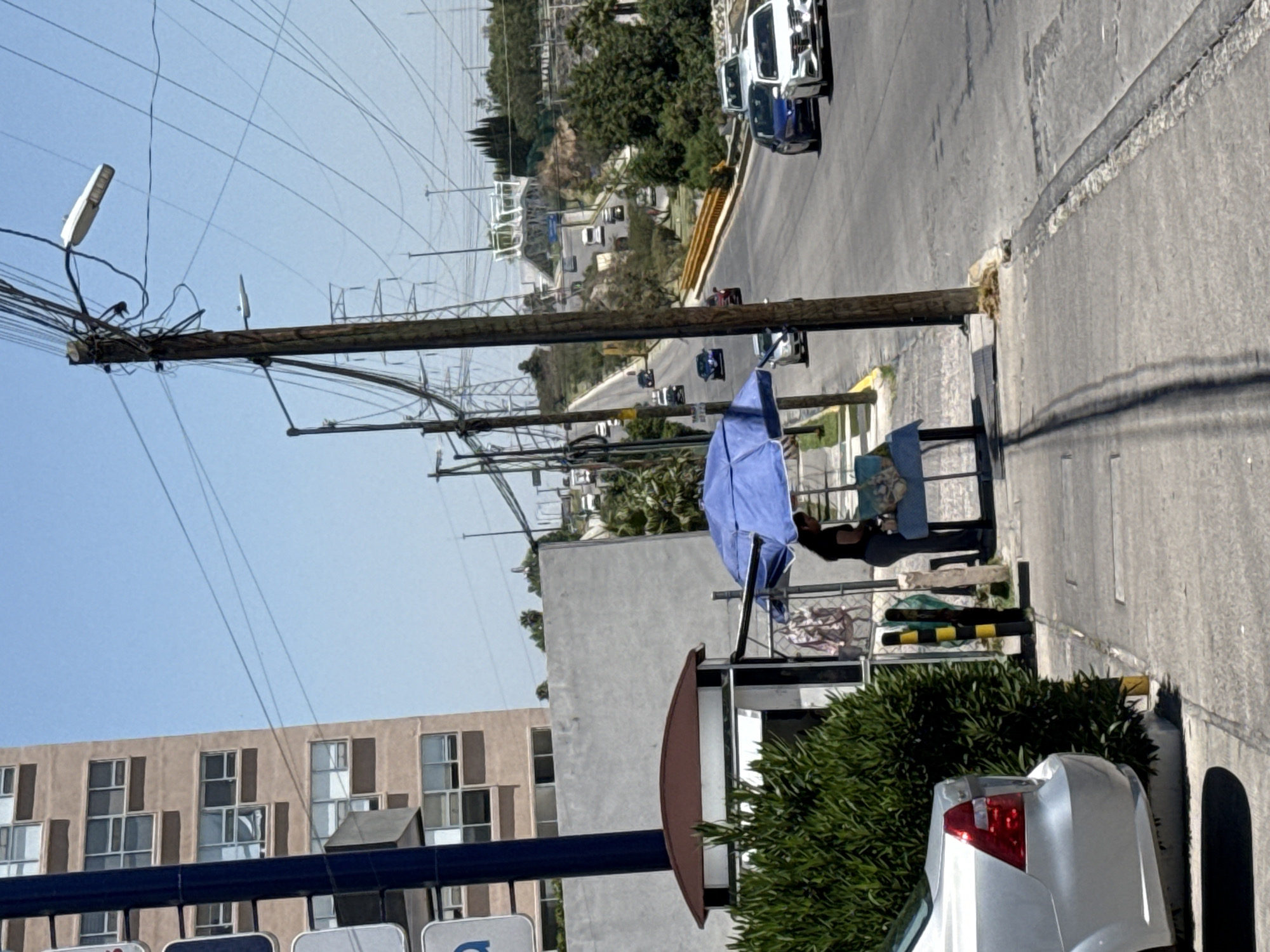 This image captures a rotated view of a vibrant urban street scene in Puebla, Mexico, under a clear, bright blue sky. The scene is characterized by a central wooden utility pole heavily adorned with a dense network of electrical wires and cables, extending vertically through the frame. Several streetlights are attached to the pole.
On the right side of the frame, alongside the street which appears to ascend diagonally, a person is seated under a large blue umbrella, suggesting a street vendor's stall or a public waiting area. A black and yellow striped barrier is visible near this setup. In the immediate foreground, the rear bumper and taillight of a silver car are visible. Further down the street, a white pickup truck and a blue car are prominent, with more vehicles receding into the distance. The white pickup truck displays the letters "NP" and "SA" on its side.
On the left, a multi-story building with numerous windows and brown shutters stands tall, adjacent to a modern concrete building with a smooth facade. The background features hills covered with trees and scattered buildings.
The scene depicts typical daily urban life, with vehicles on the street and a visible roadside presence. The abundance of overhead power lines is a notable characteristic common in cities within Mexico. The bright sunlight and distinct shadows suggest it is daytime, likely mid-morning or early afternoon. A brown, curved structure at the bottom left appears to be part of a public transport sign, with visible text fragments hinting at "BUS" or "AUTOBUS."
