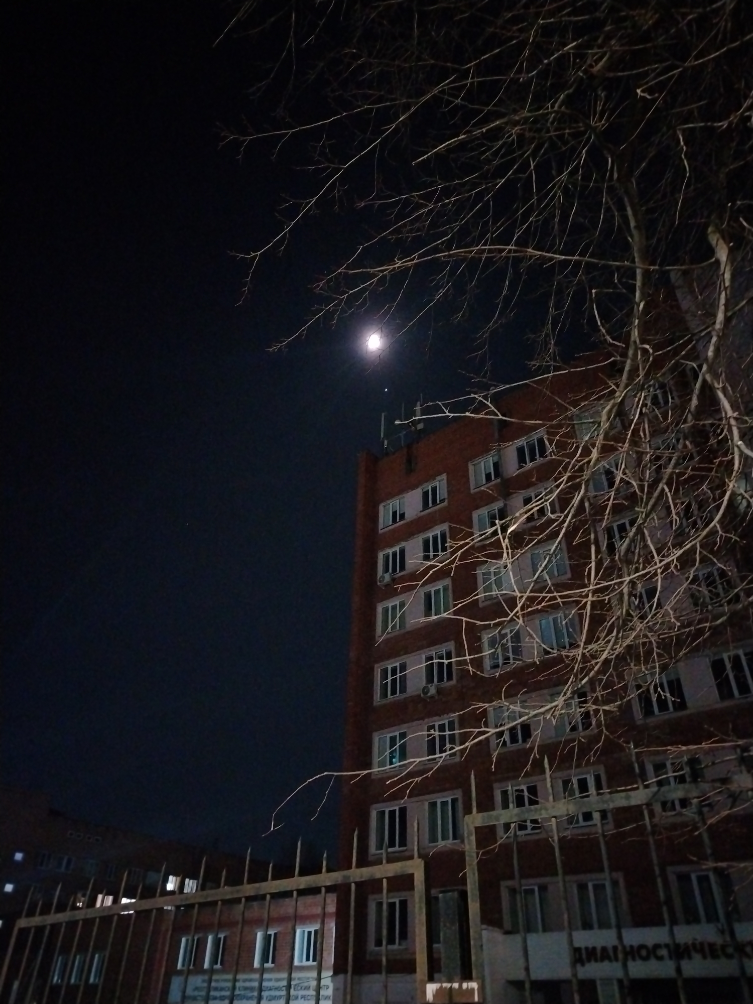 The image depicts the exterior of a multi-story brick building at night, viewed from ground level. The sky is dark, with a bright moon visible in the upper left quadrant. Bare tree branches, devoid of leaves, extend from the right and central foreground, partially obscuring the building. In the immediate foreground, a dark metal fence with vertical bars and horizontal crossbeams is visible, further obstructing the view of the lower portion of the building.
The building itself is constructed of red-brown brick and features multiple rows of rectangular windows, some of which are illuminated from within. Several white rectangular air conditioning units are affixed to the exterior walls below windows on various floors. On the upper part of the building, near the roofline, several antenna-like structures are visible against the night sky. On the lower section of the building, behind the fence, white text is visible, partially reading "ДИАГНОСТИЧЕСКИЙ" (Diagnostic).
This building is identified as part of the Republican Clinical Diagnostic Center (Республиканский клинический диагностический центр, РКДЦ) located at **Lenina Street, 81, Izhevsk, Udmurt Republic, Russia**. There are no visible people, specific actions, or interactions depicted in the scene. The overall environment is that of a city at night.