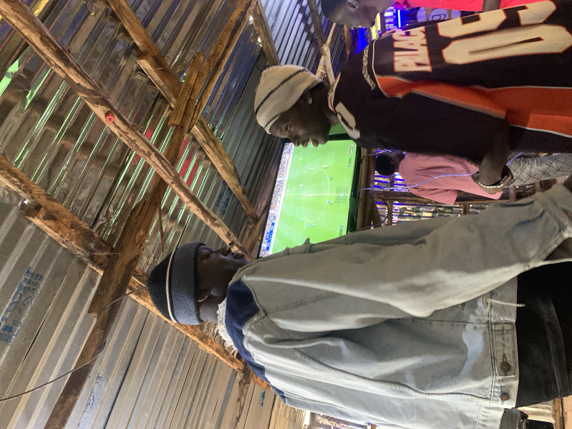 Two young men are standing and facing each other. The man on the left is wearing a light blue denim jacket and a grey beanie. The man on the right is wearing a black and orange jersey with "PALACE 05" visible on it, and a white beanie. Both men appear to be looking at a television screen in the background, which is displaying a soccer game. They are standing under a corrugated metal roof supported by wooden beams. The environment suggests an outdoor or semi-outdoor entertainment area, possibly a bar or viewing center, in Durumi, Nigeria. The lighting is dim, with the television screen providing the main light source for the immediate area. There are other people blurred in the background.
