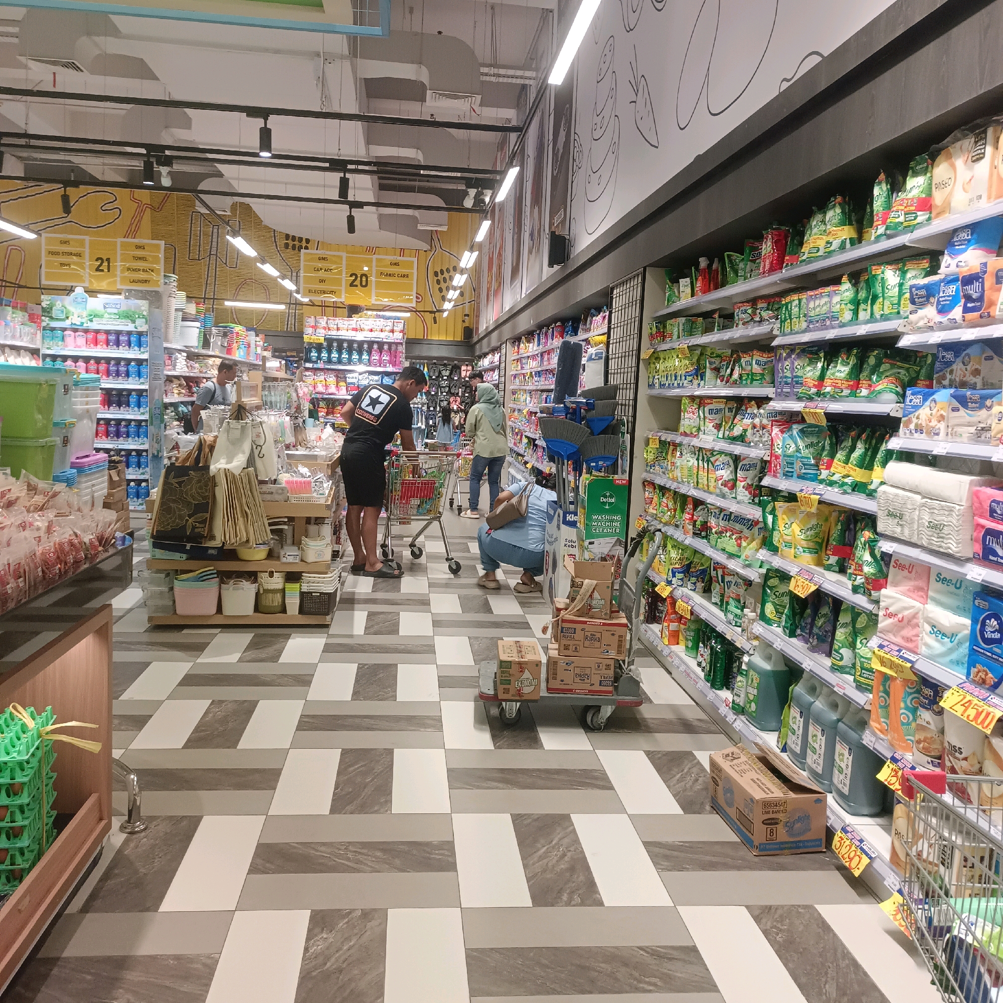 The image depicts the interior of a brightly lit supermarket in Surabaya, Indonesia. In the foreground, a person wearing a black t-shirt with a white star graphic and shorts pushes a shopping cart. To their right, another person kneels beside a stack of boxes. Behind them, a woman in a light blue hijab walks away from the camera. Shelves stocked with various consumer goods, including cleaning supplies, packaged food items, and toiletries, line the aisles. The floor features a geometric pattern of grey, white, and dark grey tiles. A prominent yellow mural with cartoonish illustrations of kitchenware and food adorns the wall in the background. Several signs, some with prices, are visible on the shelves. The overall scene captures a typical shopping experience in a modern Indonesian supermarket.