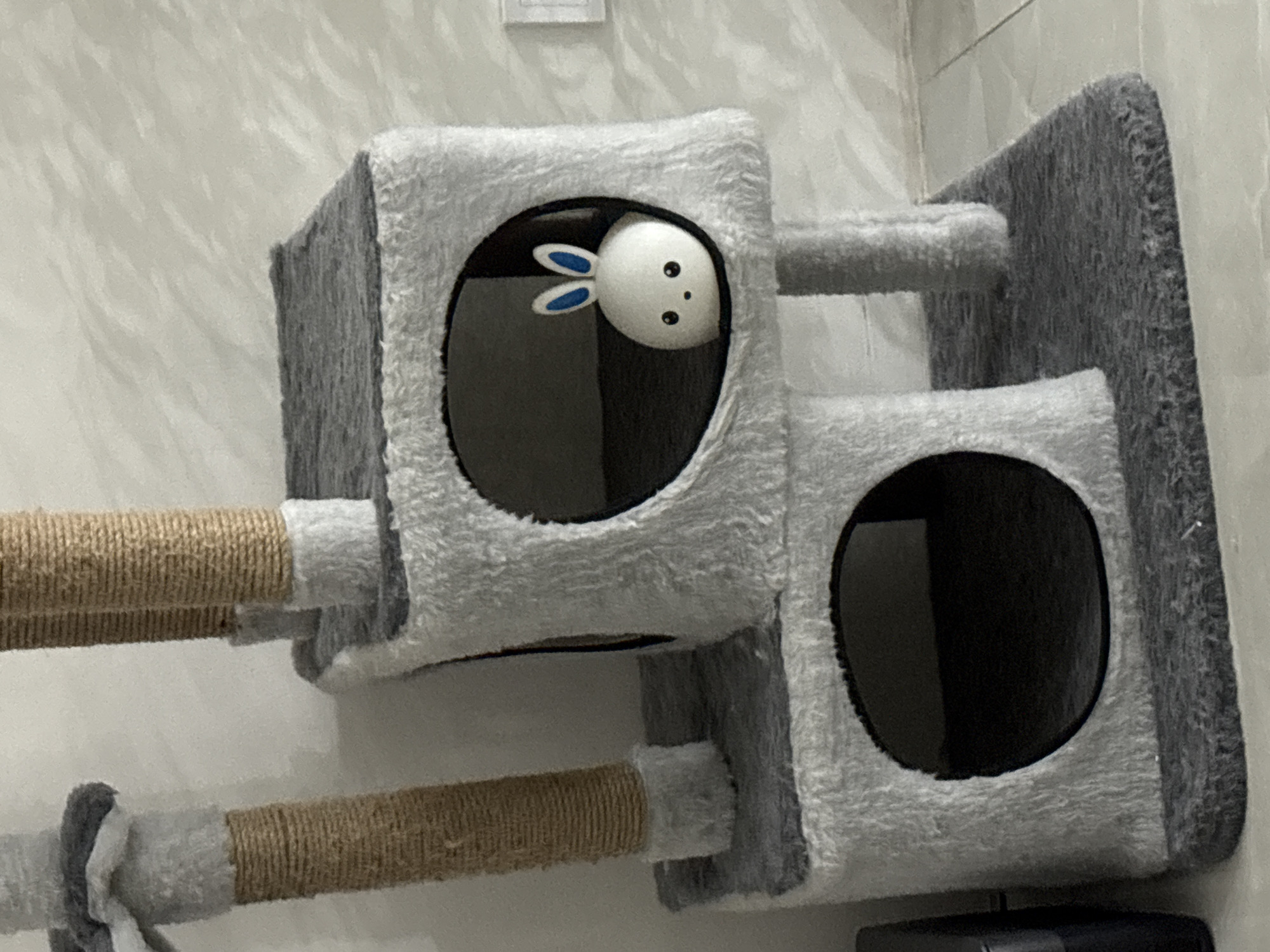 The image shows a gray cat tree with two square-shaped condos, each featuring a circular opening. The condo on the top level has a white bunny toy visible through the opening. Two sisal-wrapped scratching posts extend horizontally from the side of the structure. The entire cat tree is mounted on a wall. The location is Unknown, Unknown.