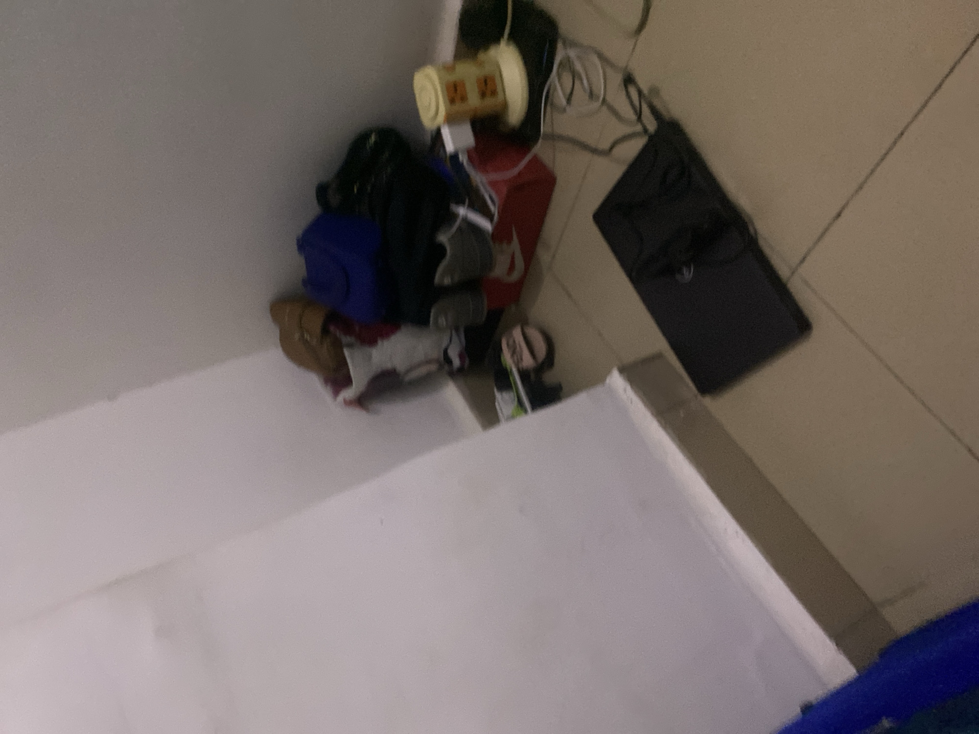 The image depicts a corner of a room with items on the floor and a wall. In the background, a pile of shoes and bags is visible, including what appears to be a blue bag, a pair of brown sandals, and several other footwear items. A red Nike shoe box is also present, with some electronic devices and cables plugged in near it. To the right, a small yellow appliance with orange detailing is positioned on a black surface. In the foreground, a black laptop is placed on the tiled floor. The walls are plain white, and the floor is covered in beige tiles. The lighting is dim, suggesting an indoor setting, likely in Howayi, Nigeria.