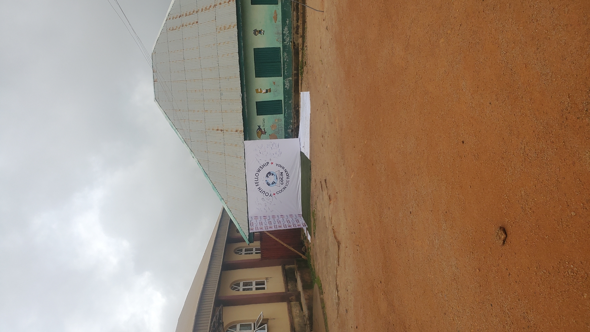 A wide-angle, eye-level shot depicts an outdoor scene in Jos, Nigeria, under an overcast sky. The foreground is a dusty, reddish-brown ground, with a few small stones scattered about. In the midground, a large white banner is propped up, featuring the text "YOUTH FELLOWSHIP COCIN LCC Tudun Wada." The banner is adorned with signatures and drawings, some of which appear to be cartoonish characters. Behind the banner, a green building with shuttered windows and some decorative art, possibly depicting cartoon characters, is visible. To the left of the green building is a cream-colored building with arched windows, resembling a church or institutional structure. A wooden fence separates the two buildings. The overall impression is of an informal gathering or event taking place in an outdoor community space.
