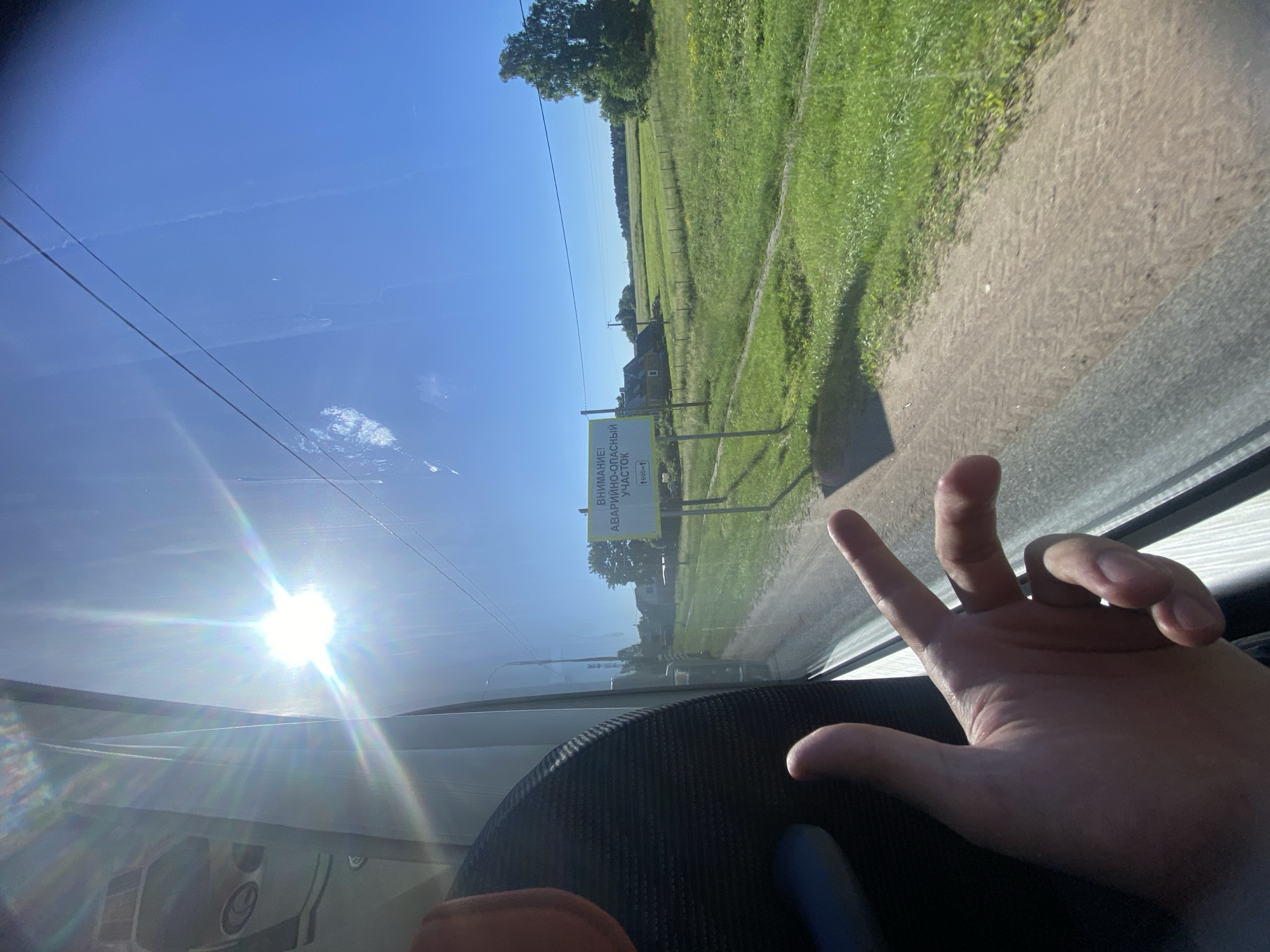 The image shows a view from inside a vehicle, looking out of a window towards a sunny rural landscape. A person's hand is visible in the foreground, gesturing with two fingers extended. Through the window, a bright sunbeam with lens flare illuminates the scene. A road or dirt path runs alongside a lush green field. In the distance, there are several houses, trees, and power lines. A yellow sign with Russian text is prominently displayed on a post, indicating an "EMERGENCY-DANGEROUS SECTION." The sky is clear and blue, with a few wispy clouds. The location is identified as Bol'shoe Teshkovo, Russia.