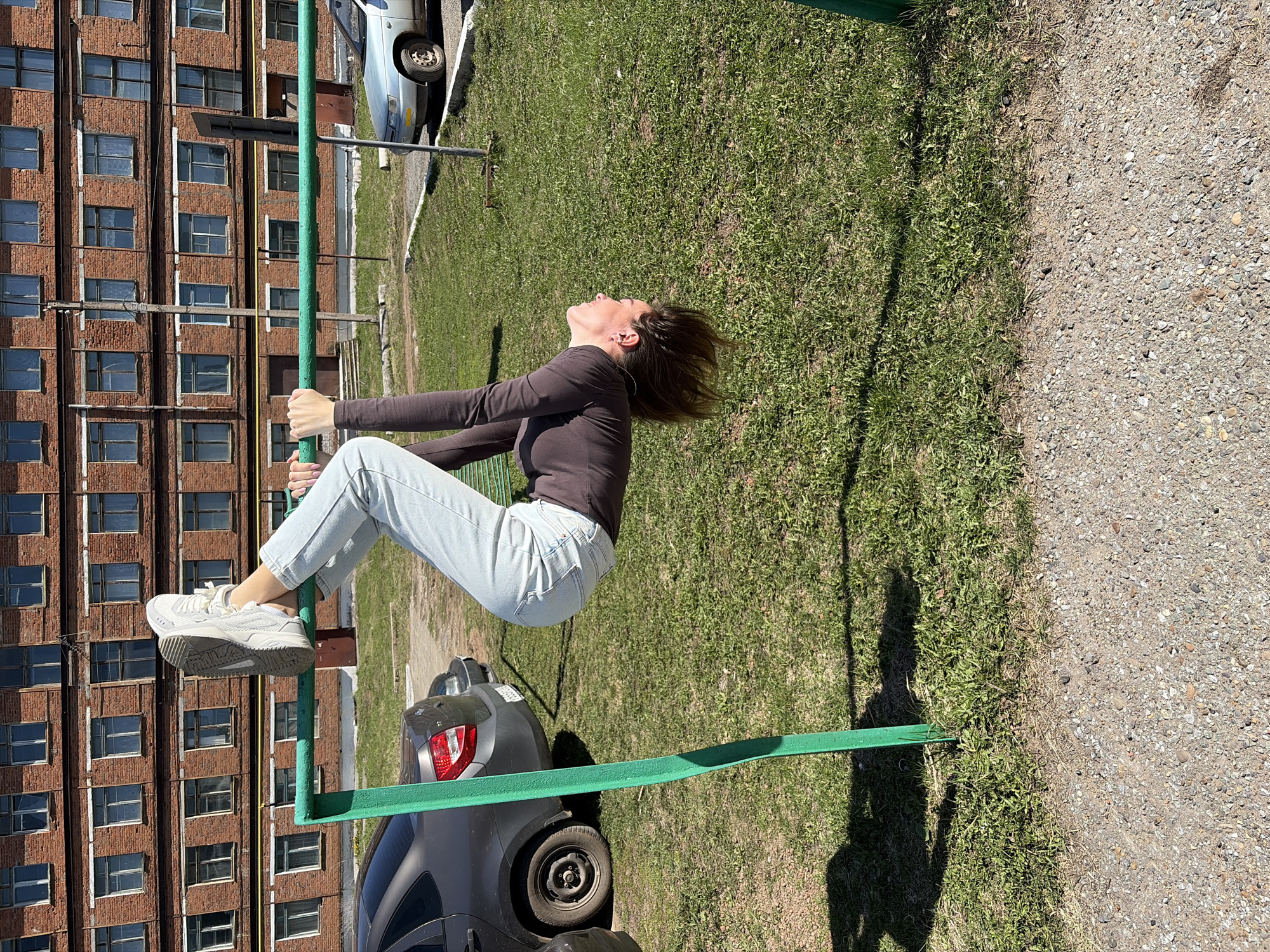 A woman hangs upside down from a green metal jungle gym. She wears a dark brown, long-sleeved shirt, light blue jeans, and white sneakers. The backdrop includes a large brick building with numerous windows, a green grassy area, and several parked cars. A gravel path borders the grass on the right side of the image. Based on the provided context, this scene is likely in the city of Neftekamsk, Russia.