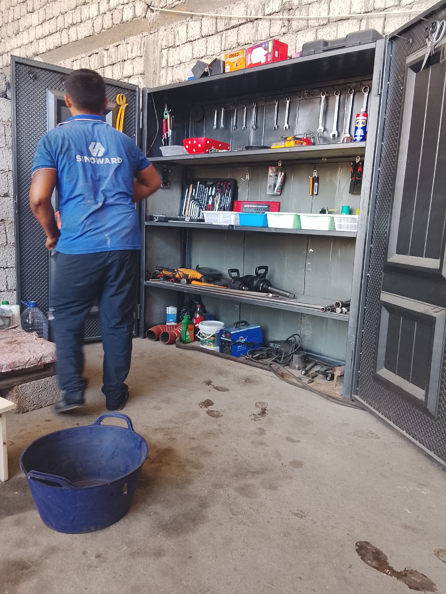 A man wearing a blue "SINOWARD" t-shirt and black pants stands in front of an open, multi-shelf metal cabinet filled with tools and equipment. The cabinet is dark gray and has two doors, one of which is fully open, revealing the organized contents. Tools such as wrenches, pliers, and other hand tools are hung on the upper shelves. Below, there are power tools, pipes, and various containers. The cabinet appears to be located in an outdoor or semi-outdoor workshop area, indicated by the concrete floor with water or oil stains, brick walls in the background, and a large blue plastic bucket in the foreground. The overall environment suggests a functional workspace. This scene is located in the city of Sabir Rakhimov, Uzbekistan.