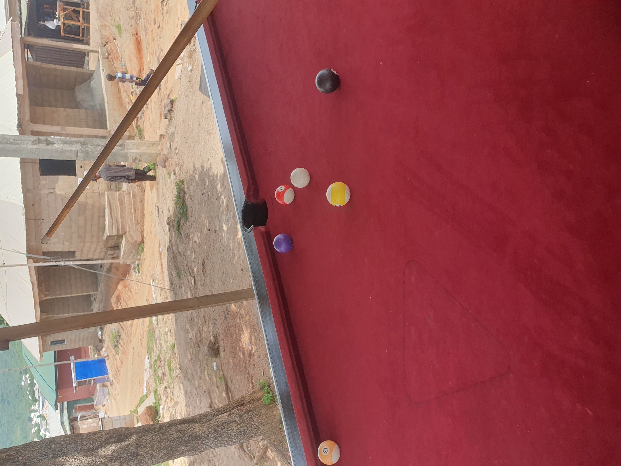 A close-up shot of a red pool table is in the foreground, with several billiard balls scattered across its surface. One ball, numbered 13 and colored orange and white, is positioned near the left edge of the table. A purple ball sits further up on the table. In the mid-ground, a group of billiard balls includes a white ball, a red and white striped ball, and a yellow and white striped ball. A solid black ball is near the right side of the table. In the background, two individuals are visible walking in a dusty, outdoor environment characterized by unfinished brick buildings, a large tree trunk on the left, and sparse vegetation. The scene appears to be in Lokoja, Nigeria, indicated by the architectural style and the general ambiance of the environment.