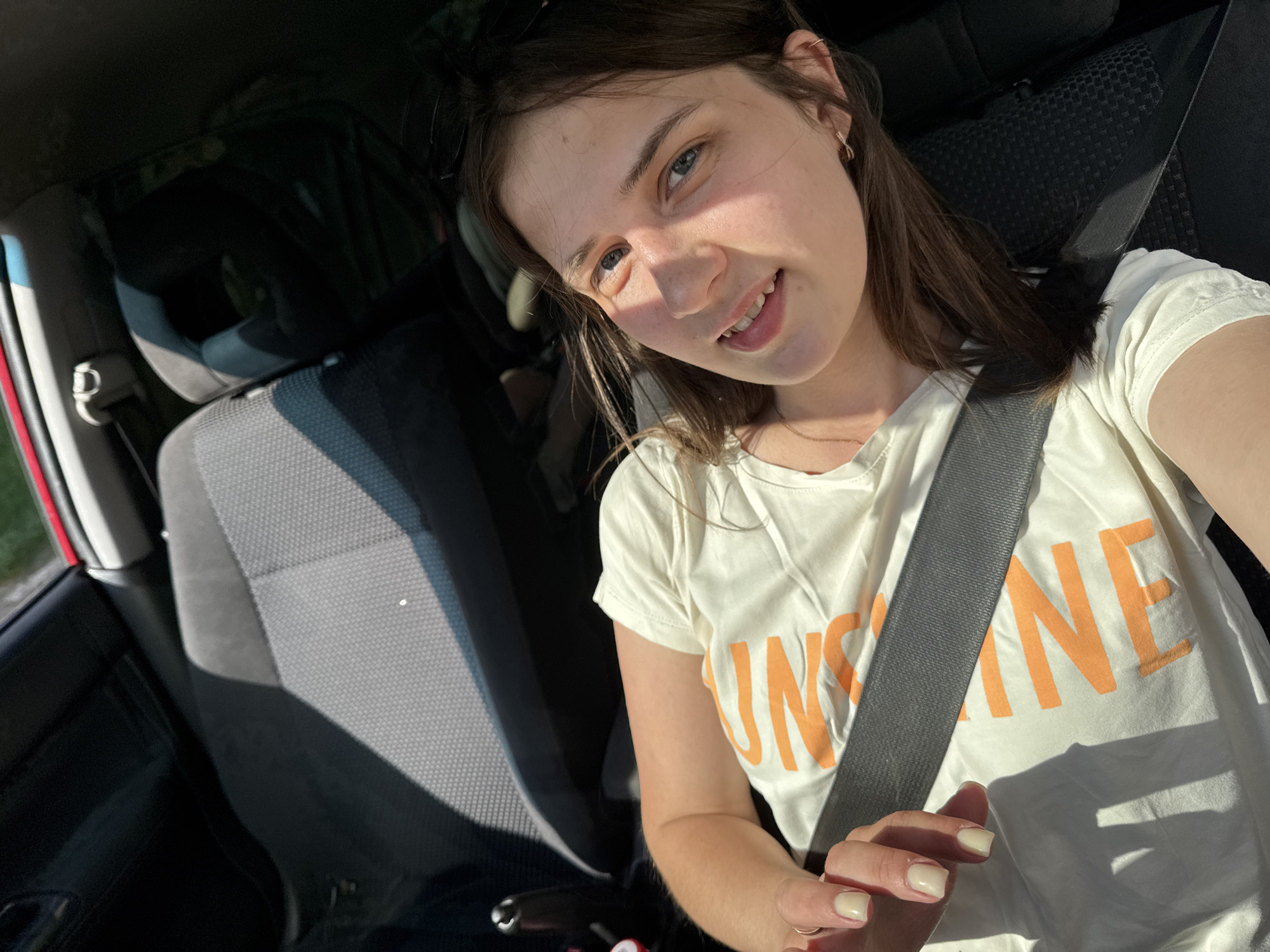A young woman is taking a selfie inside a car. She is wearing a white t-shirt with "SUNSHINE" printed in orange, and a seatbelt. The car seat has a gray fabric. The interior of the car is visible, suggesting it's either parked or stopped. The location in Chaikovsky, Russia, is not determinable from the visual information in the image.
