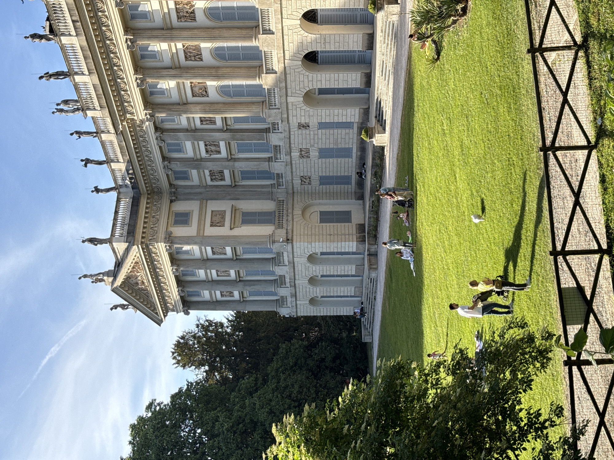 The image shows the Villa Reale in Milan, Italy. The villa is a neoclassical building with a grand facade featuring columns, statues, and decorative elements. The villa is surrounded by a lush green park with people strolling, relaxing, and interacting. A black metal fence separates the park from a path. The sky is blue with a contrail.