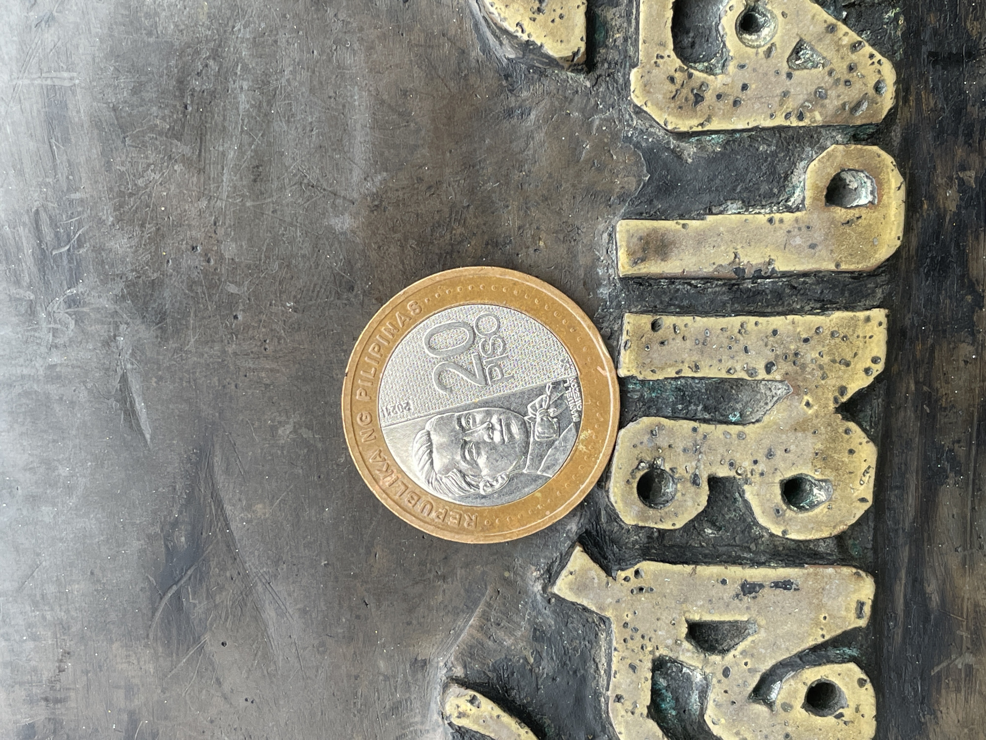 A Philippine 20 piso coin, dated 2021, featuring the portrait of Manuel L. Quezon, is placed on a weathered, textured surface with embossed characters. The surface appears to be made of a dark, possibly metal or stone material, with raised, golden-colored characters that are partially worn and corroded, showing patches of green patina. The coin is centered on the embossed characters, which are in an unknown script, possibly Thai, given the location. The overall scene suggests an outdoor environment, possibly a public monument or artwork in Na Chom Thian, Thailand, given the context provided by the prompt. The lighting is neutral, and there are no people visible in the frame.
