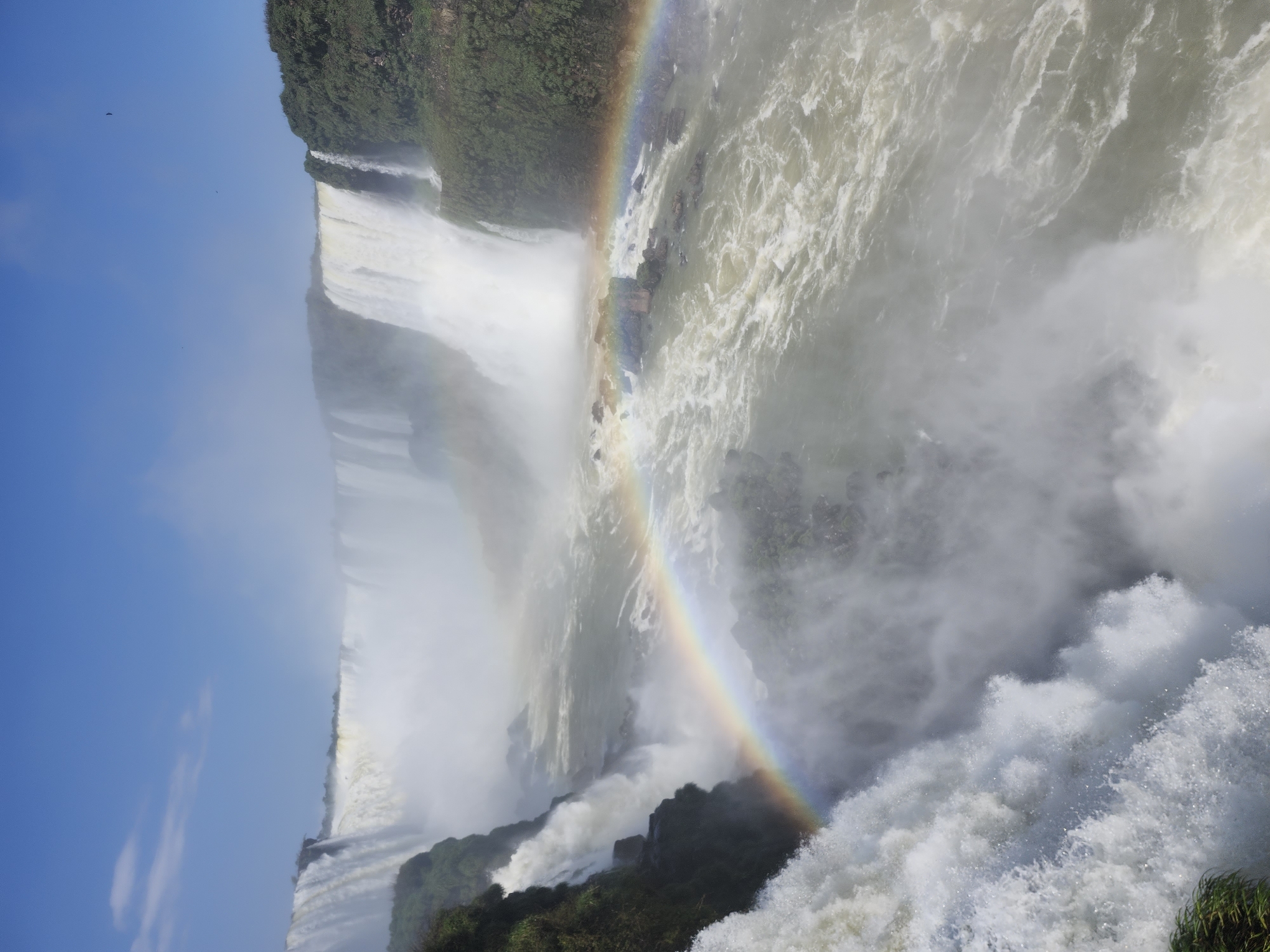 The image captures a panoramic view of the Iguaçu Falls, a majestic natural wonder located on the border between Brazil and Argentina, near the city of Foz do Iguaçu, Brazil. The falls are a system of hundreds of individual waterfalls, forming a crescent shape and creating a powerful, awe-inspiring spectacle.
In the foreground, the raw power of the water is evident as it plunges over the edge, creating a thick mist that rises into the air. The turbulent water churns and foams, displaying a range of white and grey hues. A vibrant rainbow arcs across the mist, a testament to the sunlight interacting with the water droplets. This rainbow is prominently featured, connecting different parts of the falls and adding a magical element to the scene.
The middle ground is dominated by the sheer scale of the waterfalls. Cascades of water pour down from the cliff face, creating a continuous flow that disappears into the mist below. The water appears milky white and foamy where it falls, contrasting with the darker, more serene water of the river at the base. Rocky outcrops and lush green vegetation cling to the edges of the cliffs, adding texture and depth to the landscape.
In the background, more waterfalls stretch across the horizon, their white streams blending with the soft, hazy atmosphere. The sky above is a clear, bright blue, with a few wisps of white clouds adding a touch of detail. The overall impression is one of immense natural power and beauty, with the mist and rainbow creating an ethereal and unforgettable sight.