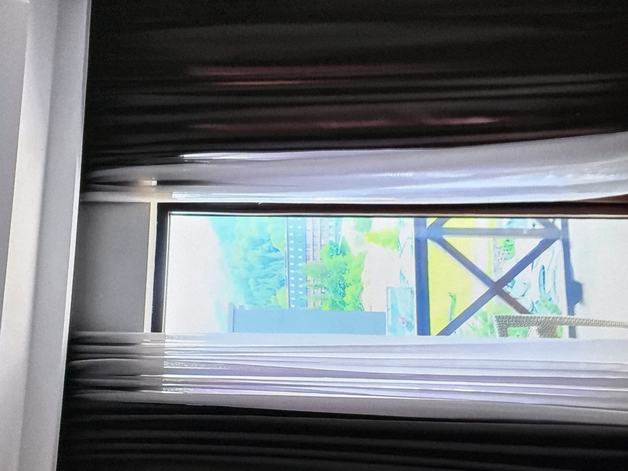 The image shows a view from a window, with dark brown curtains on the right and sheer white curtains on the left, framing a view of a building and lush green trees under a bright sky. The building appears to be multi-story with many windows and is set against a backdrop of a tree-covered mountain slope. A balcony railing with an "X" pattern is visible in the foreground, and a wicker chair sits on the balcony.