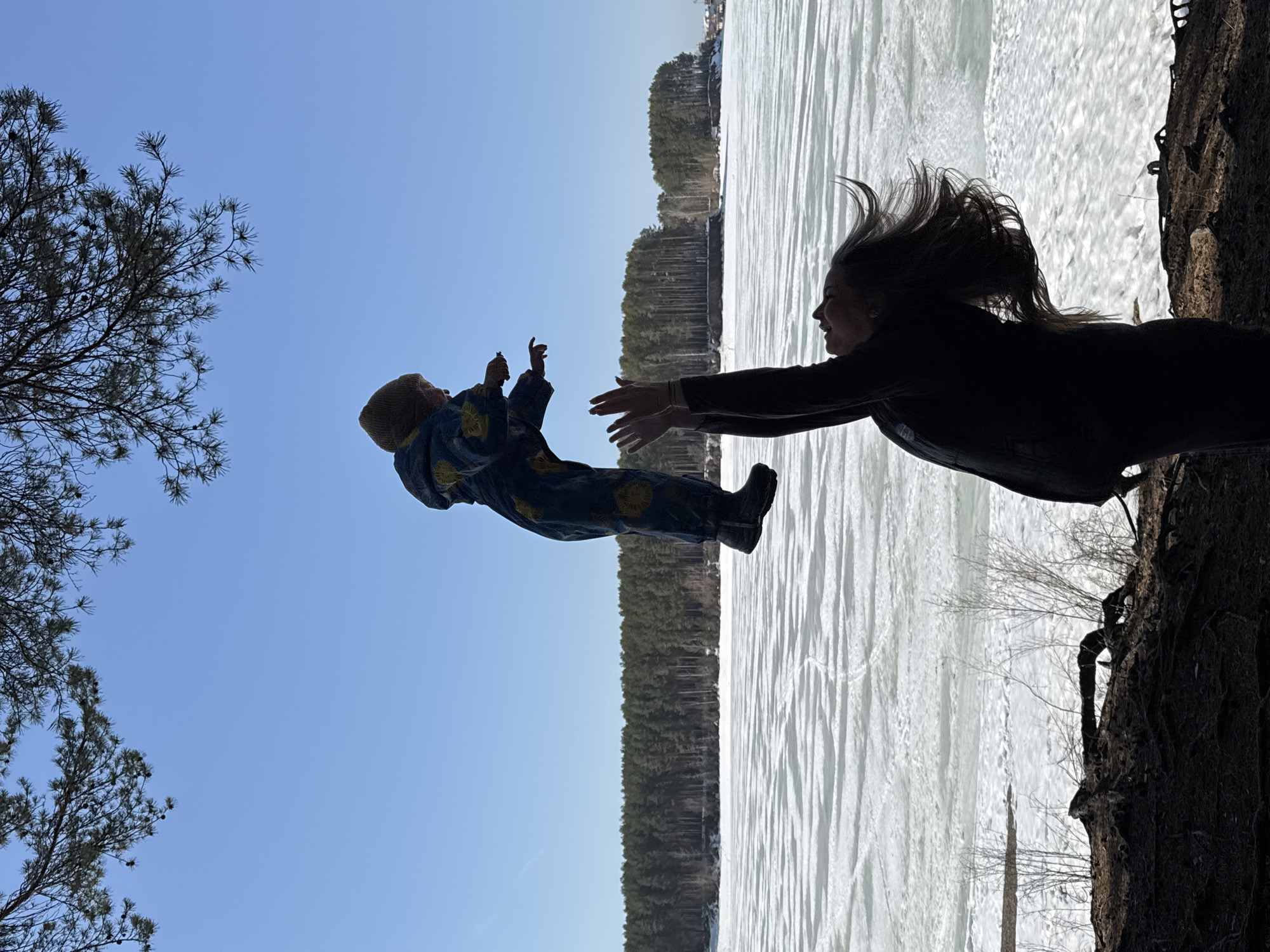 The image captures a scene in Chaikovsky, Russia, showing a woman throwing a child into the air. The woman, silhouetted against a bright sky, has long hair caught in motion. She extends her arms towards the airborne child, who is wearing a patterned snowsuit. Behind the figures is a partially frozen body of water, likely a river or lake, with a distant line of trees along the opposite bank. The foreground includes silhouetted trees and shoreline vegetation. The sky is a clear blue, visible around the trees and between the figures.