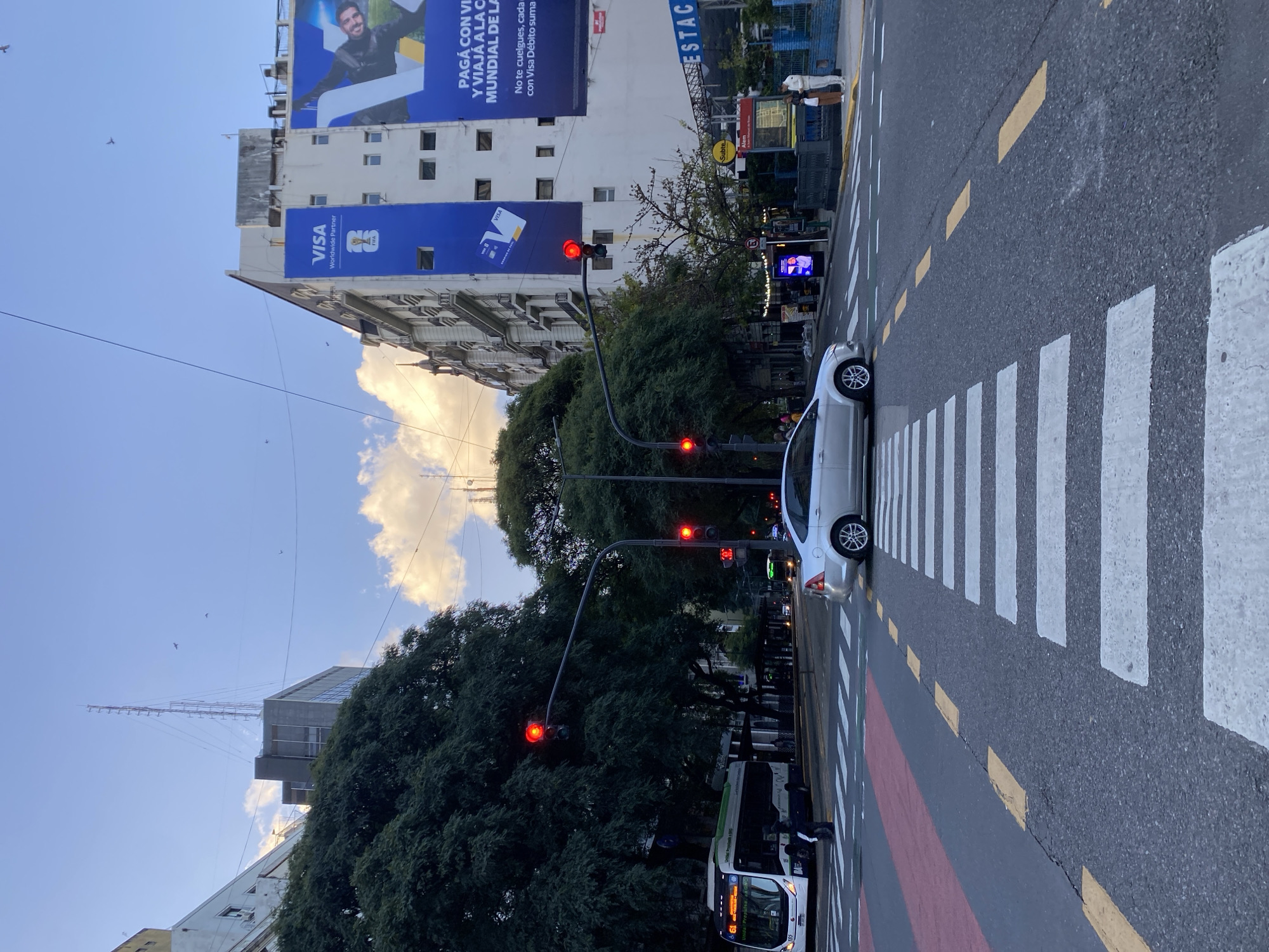 The image shows a street scene in Buenos Aires, Argentina. A white car is stopped at a crosswalk. The traffic lights show red. A bus is partially visible at the bottom of the frame. Overhead are billboards promoting VISA and featuring soccer branding. A large building with a VISA advertisement dominates the background. The sky is blue with clouds. Trees line the street. There are birds in the sky.
