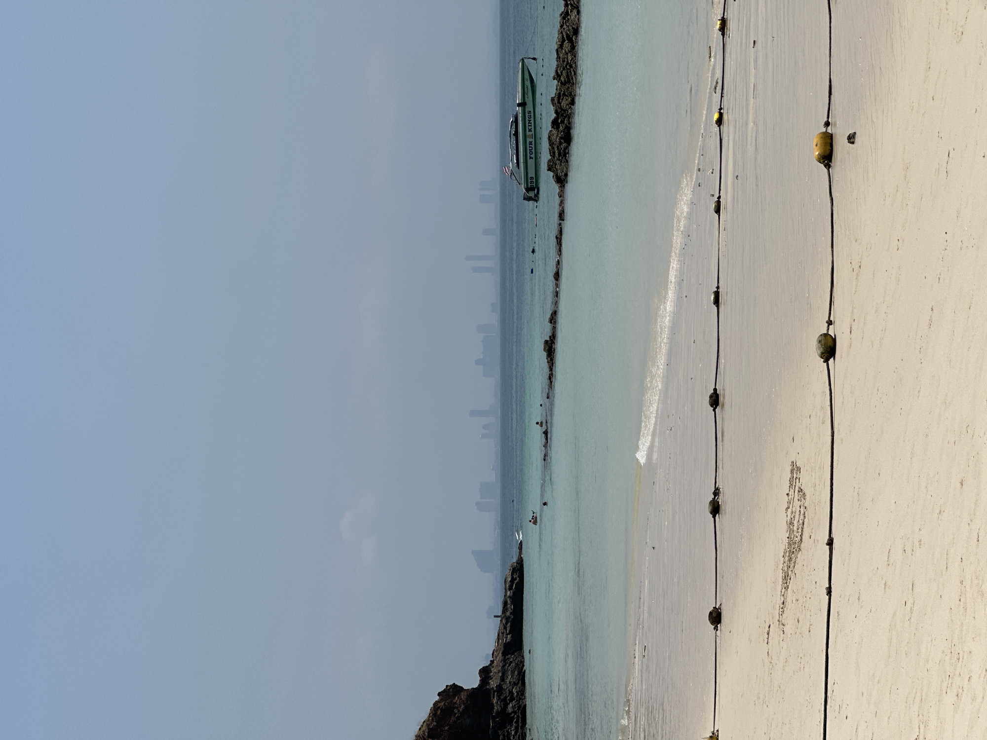 The scene is a beach in Pattaya City, Thailand, during daylight. The horizon shows a faint city skyline under a pale blue sky. Turquoise water meets a light sand beach on the right. A line of buoys runs down the beach, and a boat labeled "FOUR KINGS 839" is docked near a rocky outcropping. Other people are in the water farther along the beach.