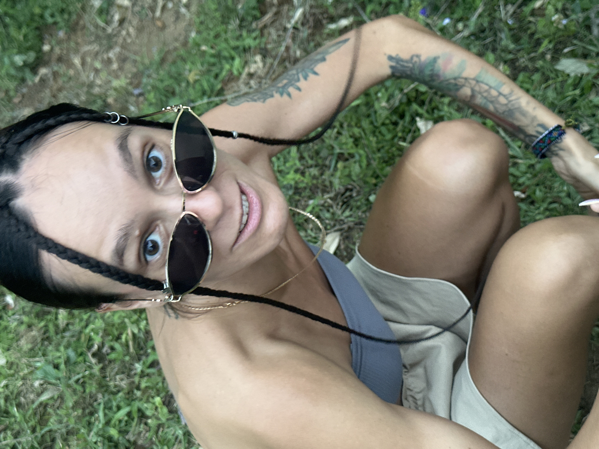 A high-angle, close-up portrait captures an individual looking directly up at the camera, seated outdoors in Nậm Dịch Commune, Vietnam. The person, appearing to be female, has dark braided hair adorned with small silver rings and is wearing aviator-style sunglasses that are slightly lowered, revealing their wide-open eyes and a gentle smile. A black cord connects the sunglasses, and large hoop earrings are visible.
The subject wears a light grey or blue top and light-colored, possibly beige, shorts. Both arms are visible and feature distinct tattoos. A colorful woven bracelet is worn on the right wrist. The setting is natural, with green grass covering the ground, suggesting a relaxed outdoor environment during the daytime. The overall impression is casual and engaging. No visible text is present in the image.
