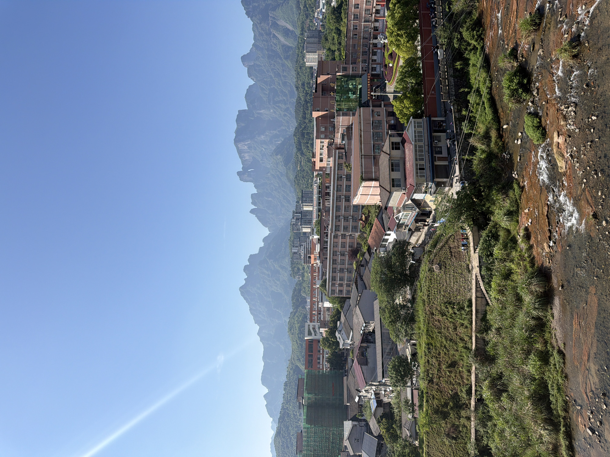 The image depicts a scenic view of Zhangjiajie, China, showcasing a blend of natural landscapes and urban development. In the background, a mountain range with distinctive rocky peaks dominates the horizon, partially shrouded in a light mist or haze, adding depth and contrast to the scene. Adjacent to the mountains, a dense cluster of buildings rises, indicating a populated urban area. The architecture includes a mix of residential and commercial structures, with varying heights and designs, reflecting the city’s growth. In the foreground, a small river or stream flows, its water reflecting the sunlight. The immediate banks are covered with vegetation, creating a natural border between the water and the built environment. A path, likely a pedestrian walkway, runs alongside the river, suggesting accessibility for locals and visitors. The sky is a clear, bright blue, providing a vivid backdrop that enhances the colors and details of the urban and natural elements in the scene.